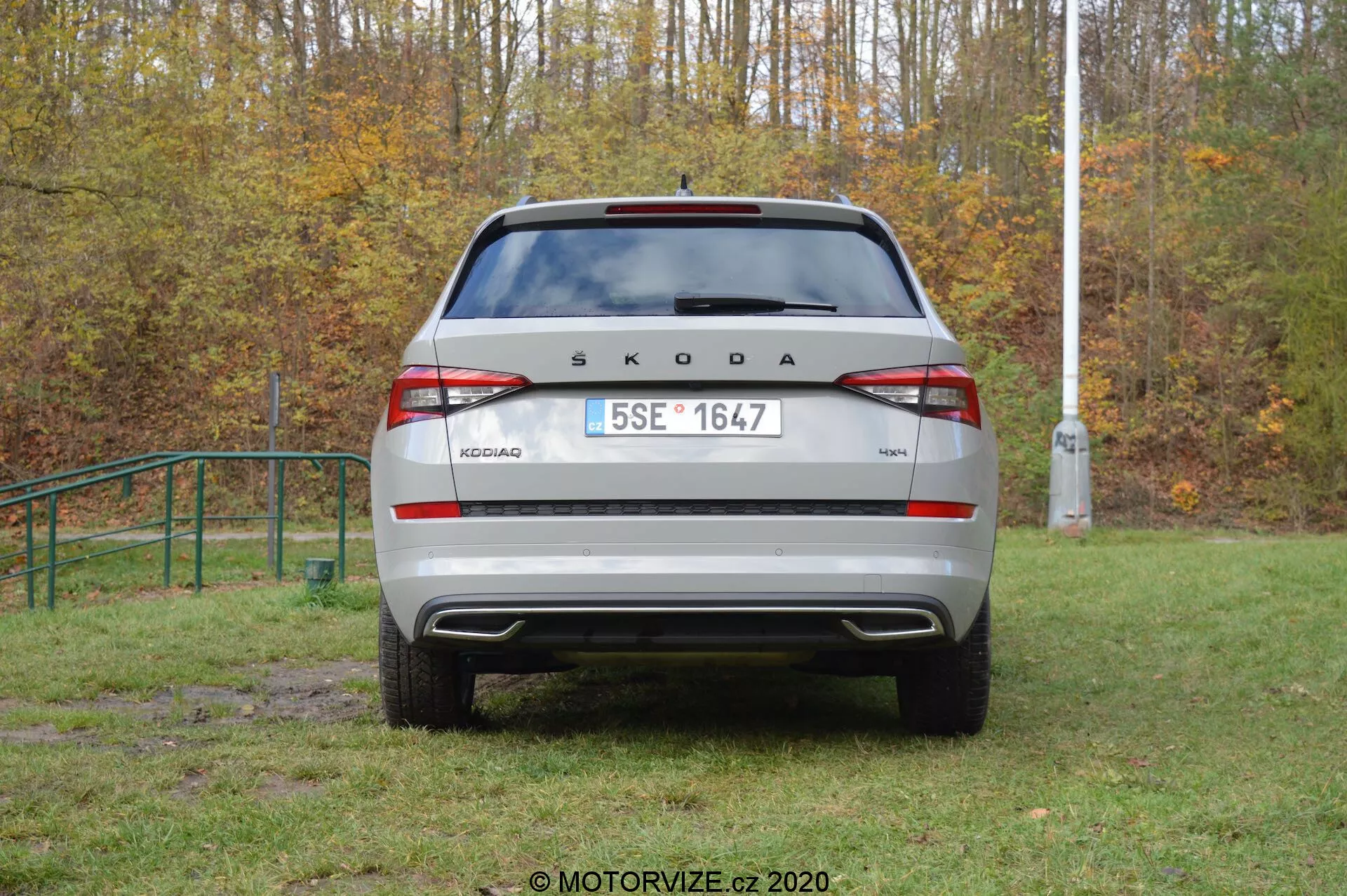 Zadní pohled na vozidlo Škoda Kodiaq 2017 ve venkovním prostředí. Značka 'ŠKODA' a model 'KODIAQ' jsou výrazně zobrazeny na víku kufru. Evropská registrační značka, moderní zadní světla s integrovanými mlhovými a couvacími světly, viditelný zadní nárazník s parkovacími senzory a zadní okno s stěračem jsou jasně viditelné. Reflexní prvky na spodní části nárazníku zvyšují viditelnost. Auto je zaparkováno na trávě, lehce blátivé oblasti, což naznačuje terénní schopnost.