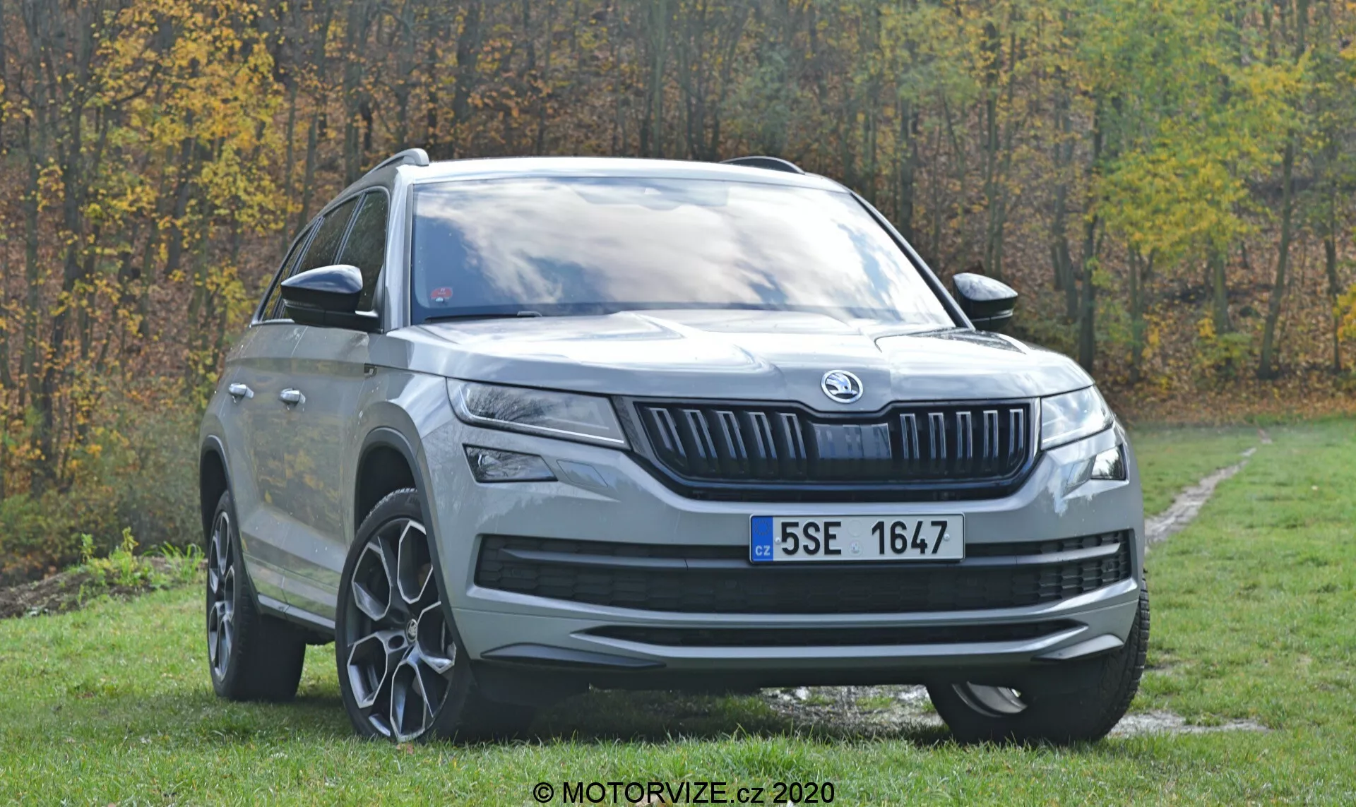 Tříčtvrteční přední pohled na moderní SUV Skoda Kodiaq 2017 zaparkované na travnaté ploše s lesem v pozadí. Viditelné funkce zahrnují výraznou přední masku s vertikálními lamelami a centrálně umístěným logem značky, elegantní horizontálně orientované světlomety s LED denními světly, vysokou světlou výšku, velká vícepaprsková litá kola, výrazné oblouky kol, stříbrný ochranný kryt, výrazné linie kapoty, karoserií zbarvená zpětná zrcátka s integrovanými směrovkami, zatmavená boční okna, mírně skloněnou střechu směrem dozadu s střešními lištami.