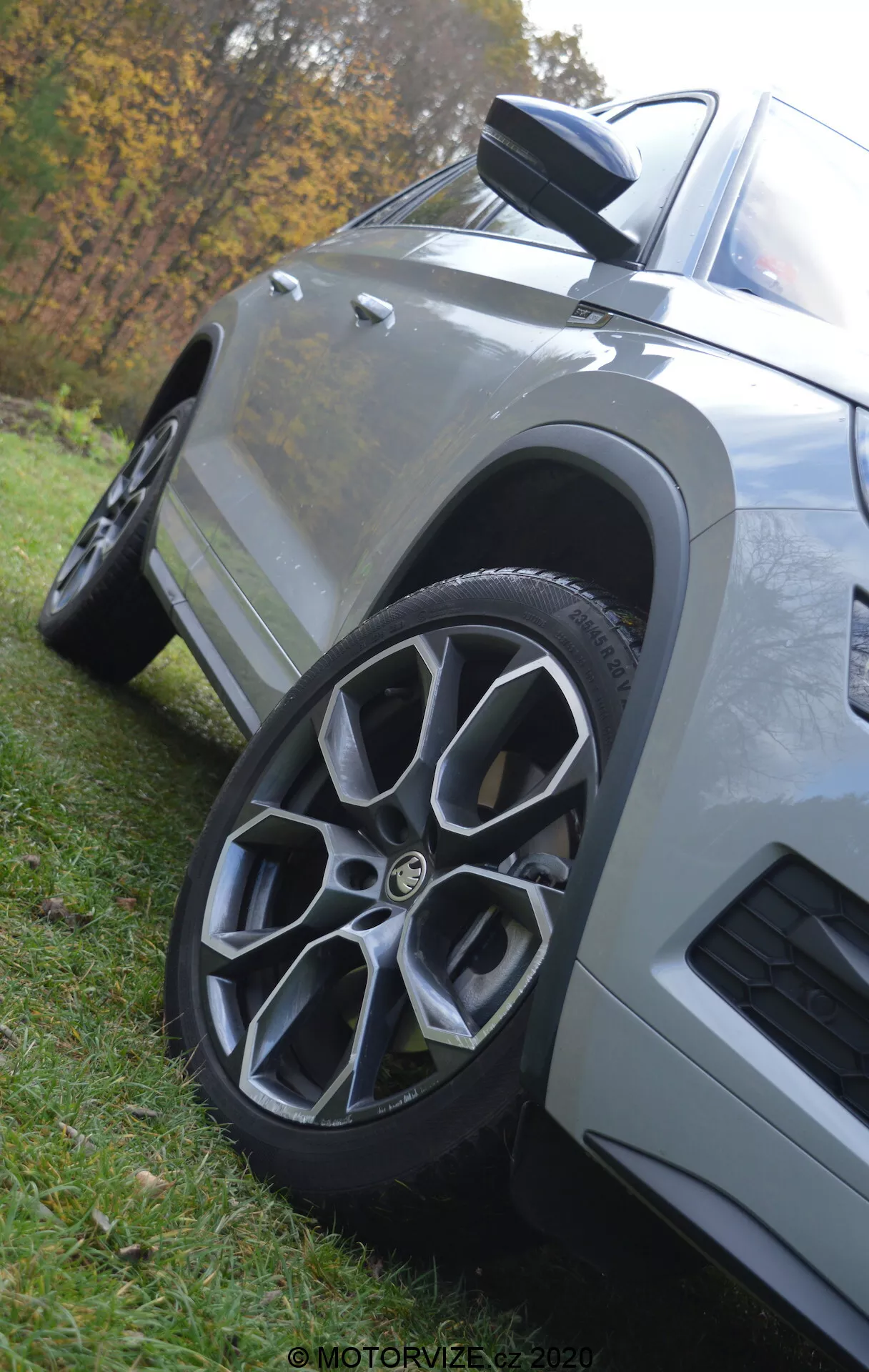 Detailní záběr z nízkého úhlu na vůz Škoda Kodiaq 2017, zvýrazňující přední kolo a blatník. Auto, zaparkované na trávě, má vícepaprskové slitinové kolo, aerodynamické boční zrcátko s integrovaným indikátorem, hranaté karosářské panely s metalickým lakem a ventilační otvor na předním blatníku, vše na rozmazaném přírodním pozadí.