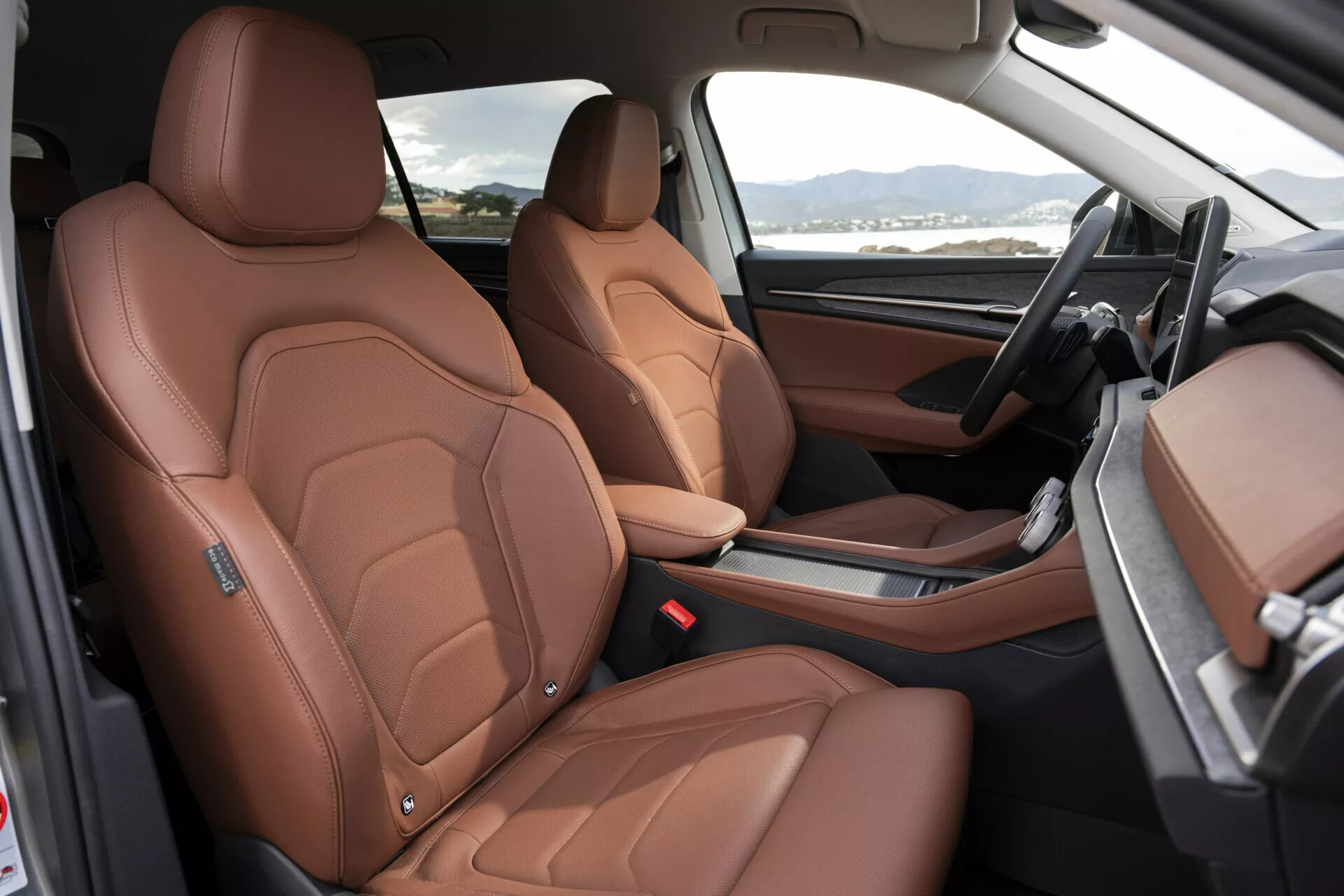 Interiér Škoda Kodiaq 2024 s luxusními předními sedadly potaženými hnědou kůží s detailním prošíváním, elektrickým nastavením sedadel a pohledem na prvky vysokého standardu jako jsou sportovně inspirované boční opěrky, černo-hnědý dvoubarevný palubní deska a minimalistická středová konzole s automatickou volící pákou, z pohledu otevřených dveří na straně spolujezdce