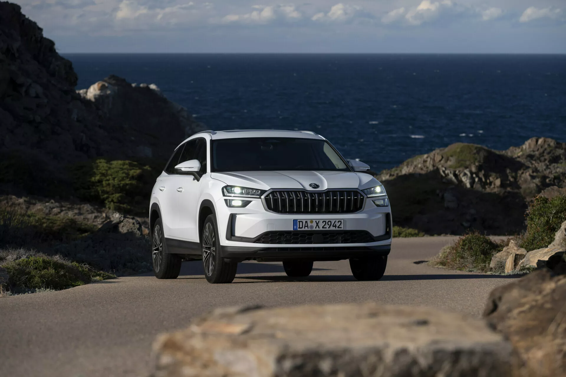 Bílé SUV Skoda Kodiaq 2024 zaparkované na pobřežní silnici s oceánem na pozadí, vyznačující se výraznou přední mřížkou s vertikálními slatami podobnými chromu, LED denními světly, slitinovými koly s více paprsky a detailním pohledem z předního tříčtvrtě úhlu