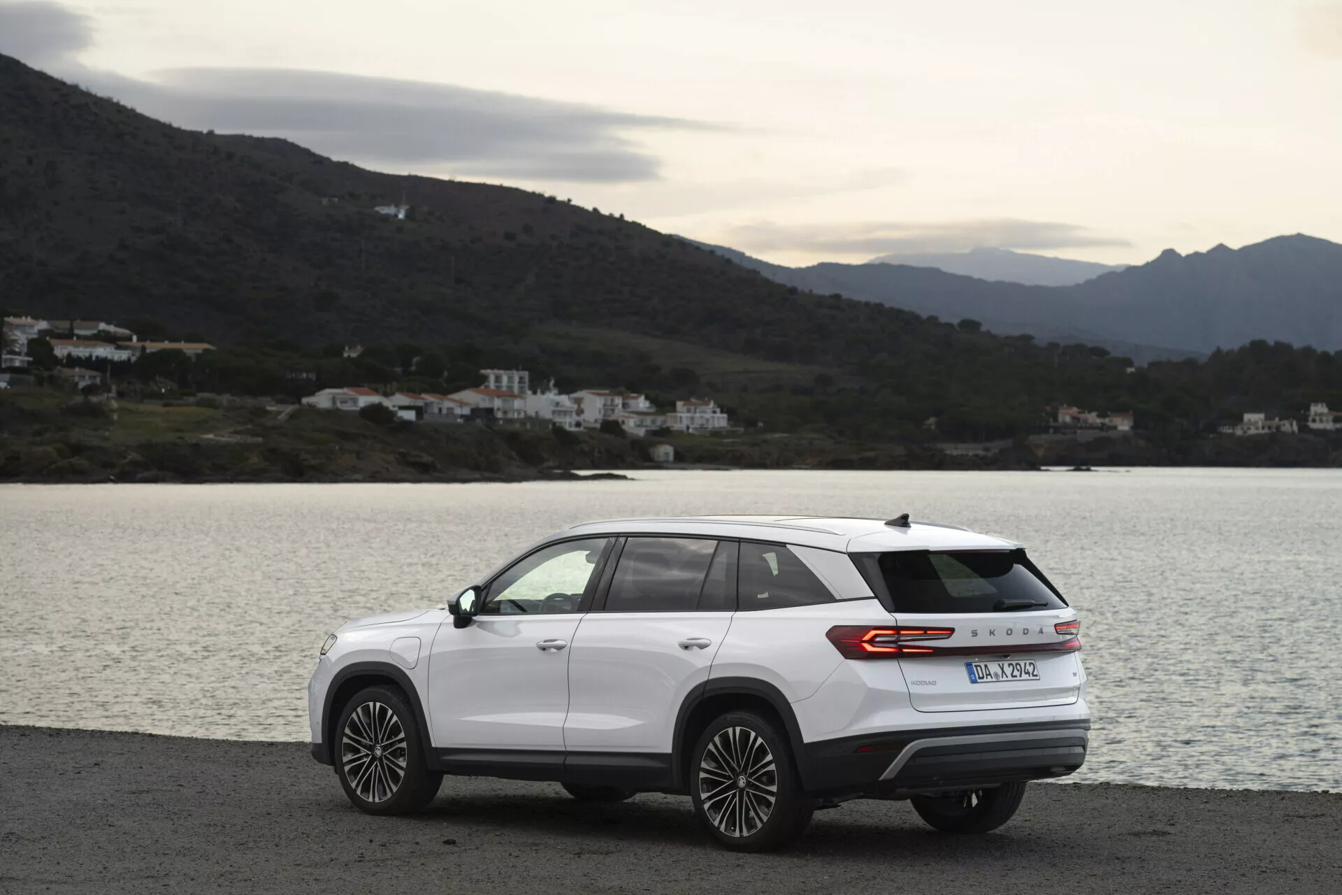 Moderní SUV Skoda Kodiaq 2024 parkované bočně na malebné pobřežní silnici, s aerodynamickým designem, pruhem zadních světel a koly z lehké slitiny s vícepaprskovým designem, na zatažený den s rozptýleným přirozeným osvětlením a s výhledem na vodu, kopce a budovy