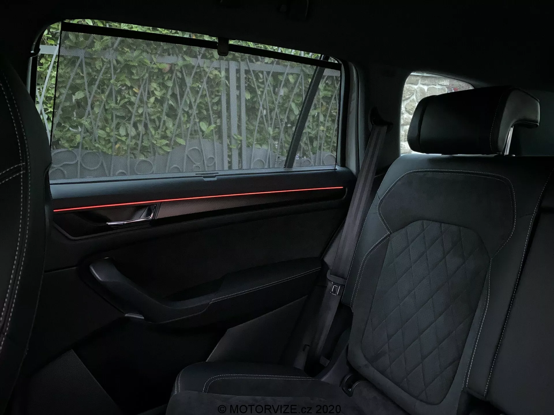Interiér vozidla Skoda Kodiaq 2017 zaměřený na zadní prostor pro cestující, zobrazující luxusní prvky jako prošívané čalounění z kůže na předních i zadních sedadlech, červený ambientní osvětlený proužek na dobře vybaveném polstrovaném panelu dveří s prémiovým obložením a částečně průhlednou sluneční clonou na okně. Důraz je kladen na pohodlí a prvotřídní designové prvky.