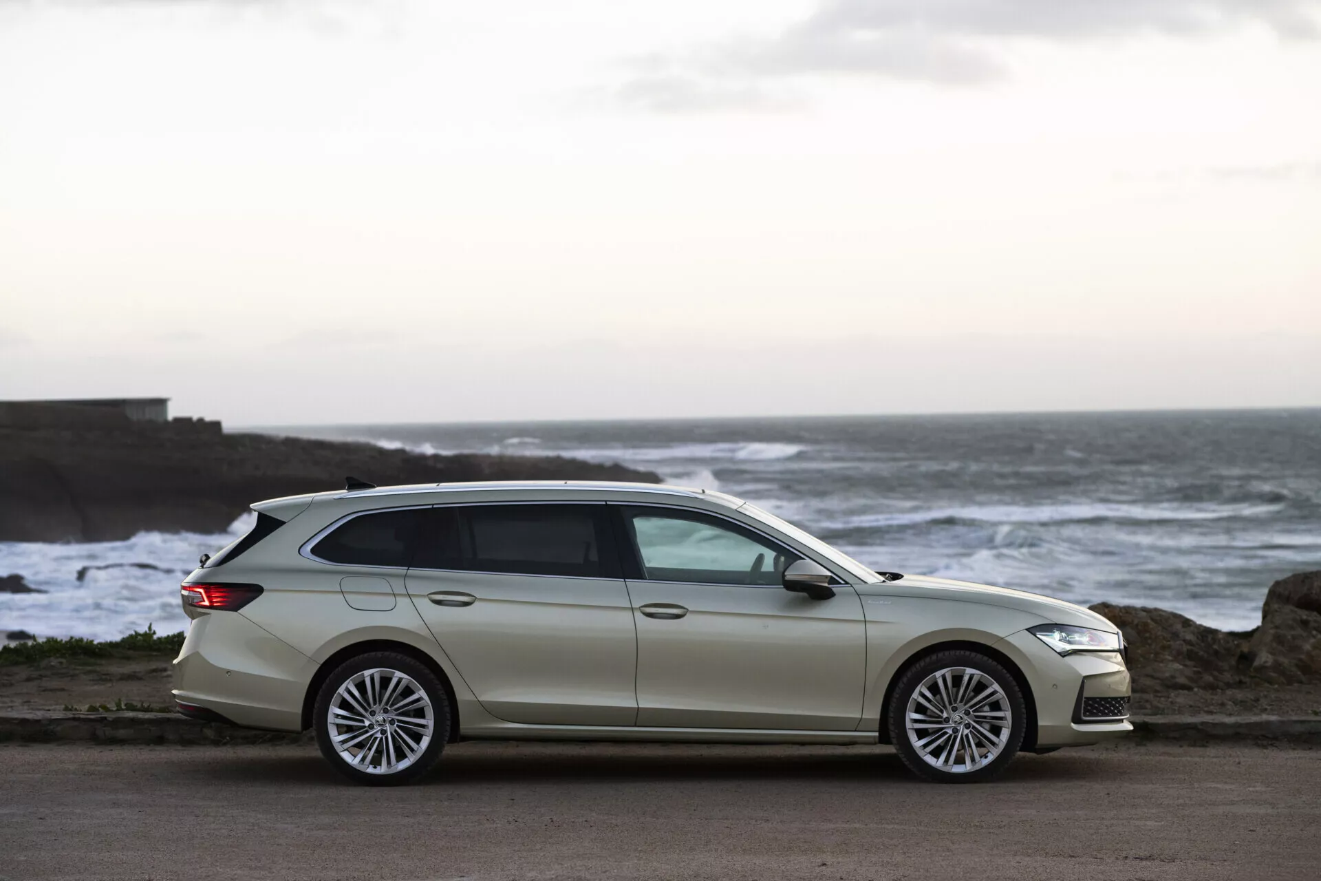 Skoda Superb Combi 2024 v světlém odstínu zaparkovaná u pobřeží v soumraku, zdůrazňující její moderní design, aerodynamickou karoserii a prémiová vícepaprsková kola z lehké slitiny