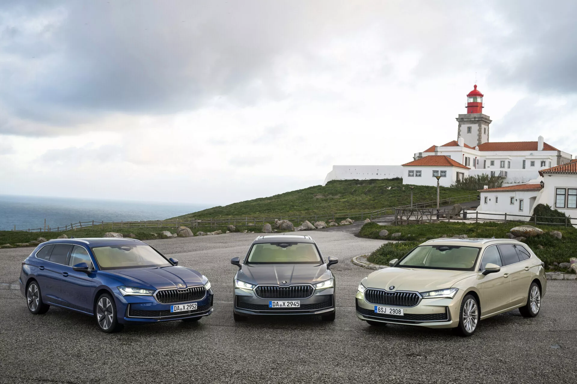 Modely Škoda Superb Combi 2024 v tmavě modré, metalické šedé a světle zlaté barvě parkují vedle sebe, předvádějící LED světlomety, chromové mřížky a kola z lehkých slitin s majákem a budovou v pozadí pod mrakem oblohou