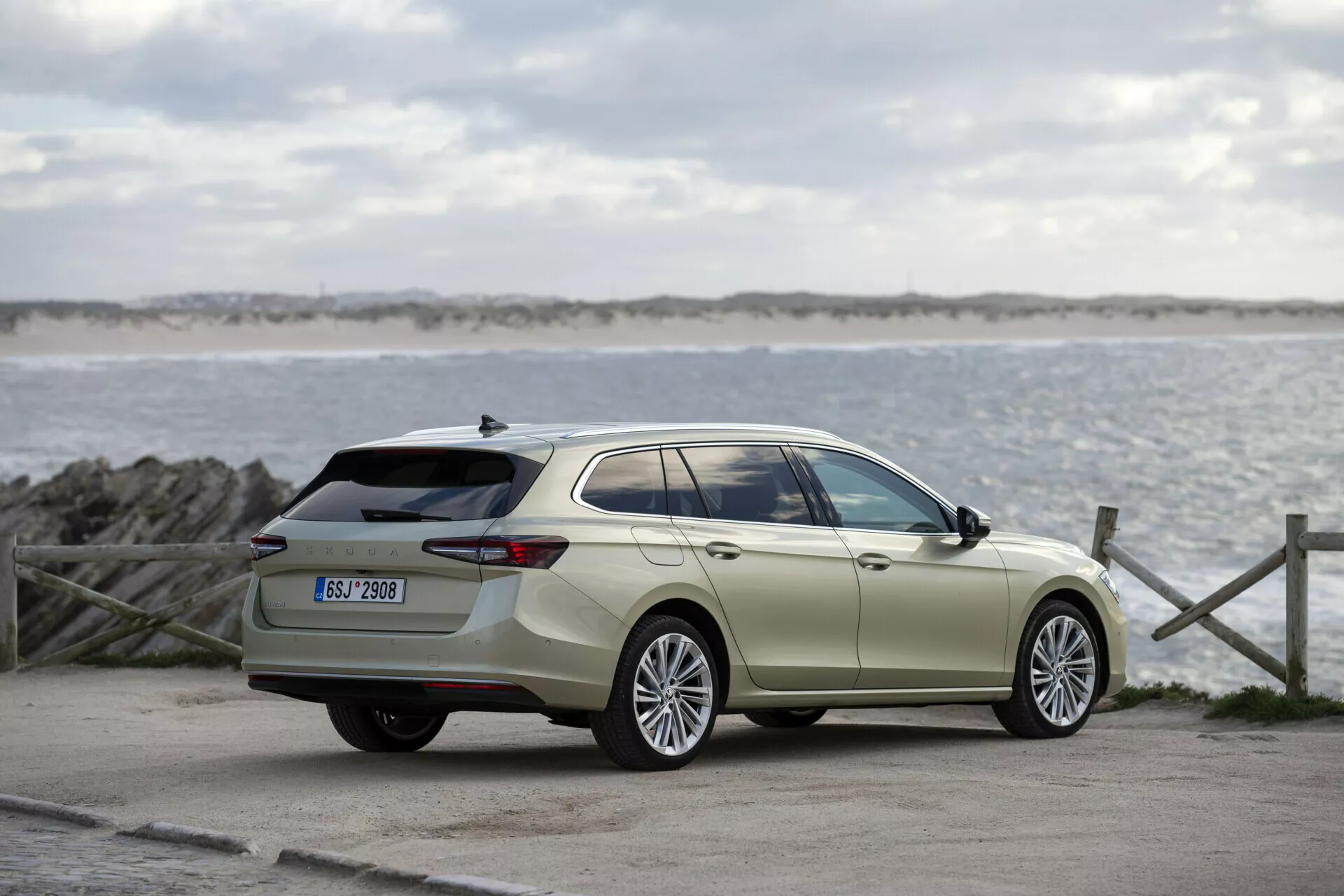 Skoda Superb Combi 2024 v exteriéru s výhledem na moře a oblohu, zobrazující plný boční profil, velká slitinová kola, dveřní kličky a zrcátka v barvě karoserie, chromové lemování oken, prodlouženou střechu, aerodynamické kontury karoserie a střešní ližiny