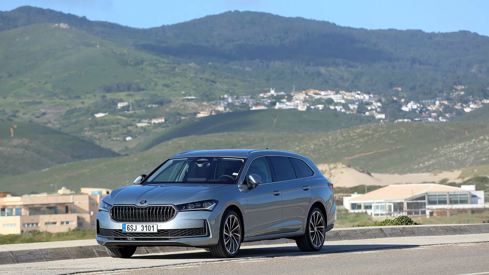 Škoda Superb Combi 2024 ve tříčtvrtečním čelním pohledu na silnici s pozadím kopcovité krajiny, vyznačující se výraznou maskou chladiče, LED světlomety a panoramatickou střechou
