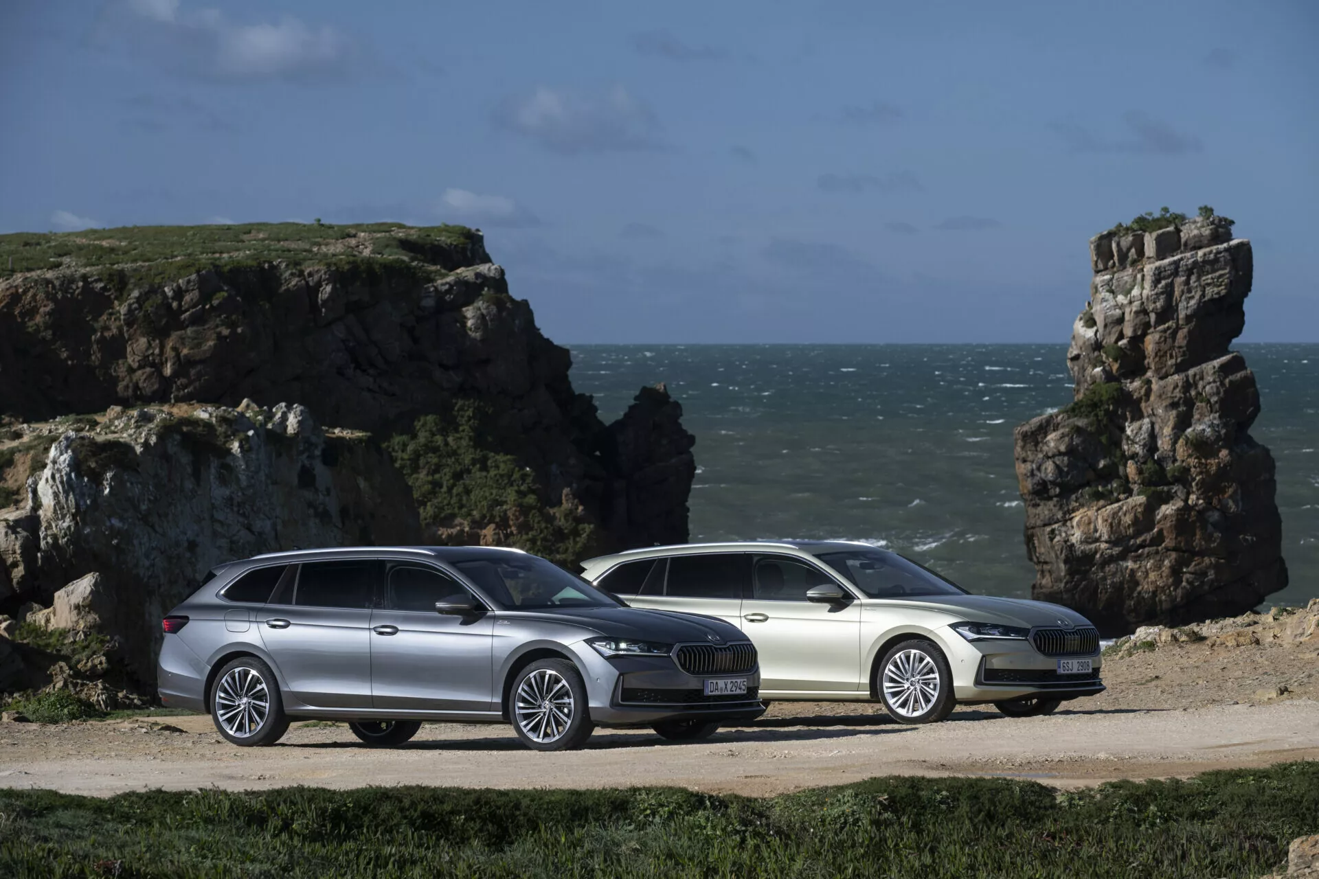 Stříbrné a bílé kombi Škoda Superb 2024 zobrazené vedle sebe ve tříčtvrtečním pohledu ukazujícím jejich aerodynamické profily, slitinová kola a integrované střešní nosiče, na pozadí pobřežní krajiny s kamennými výběžky.