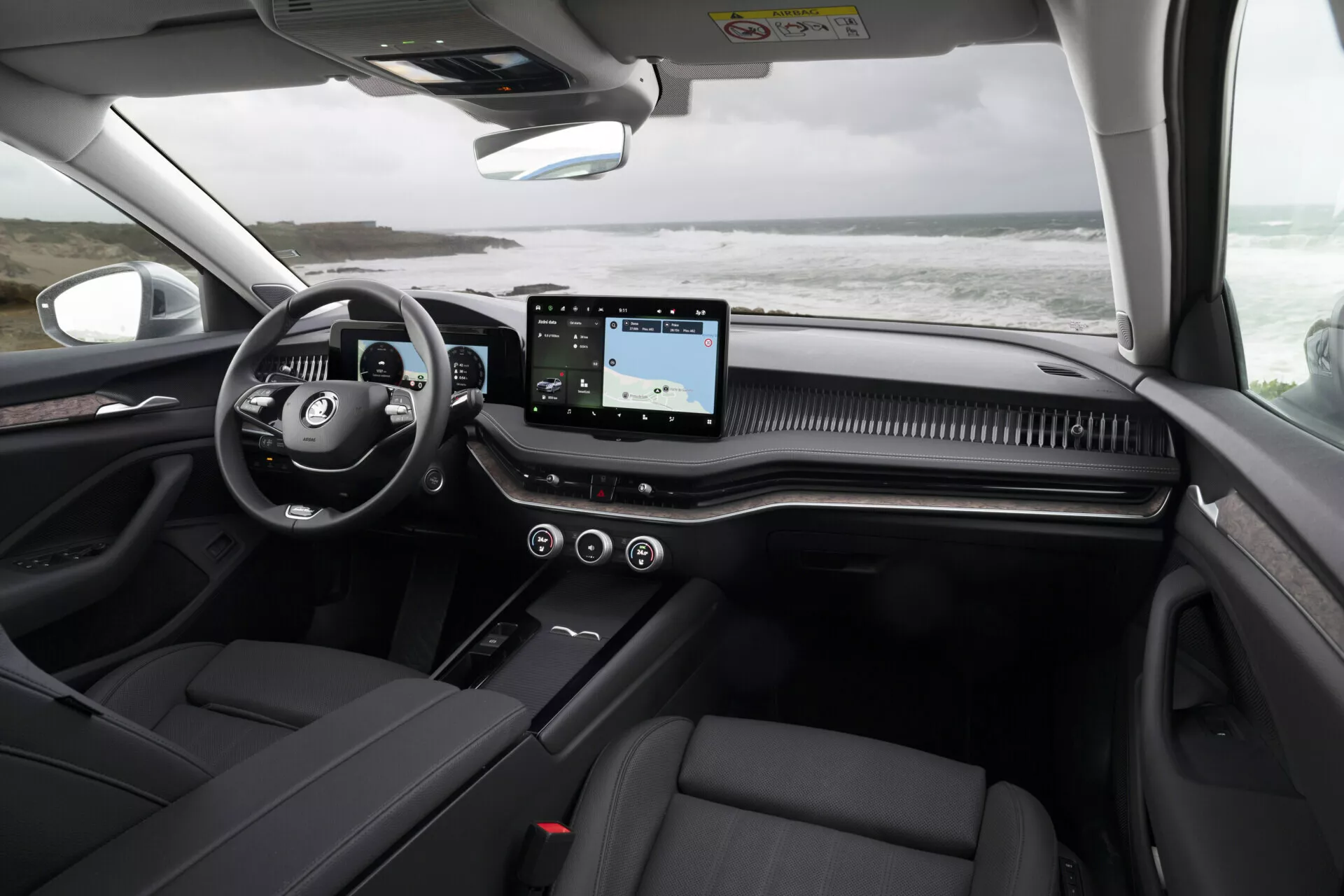 Interiér Škoda Superb Combi 2024 ukazující palubní desku z perspektivy zadního sedadla, zdůraznění multifunkčního volantu, vodorovného dotykového informačního systému, dekorativního lemu palubní desky, sedadel z tmavé kůže a detailů panoramatické střechy.