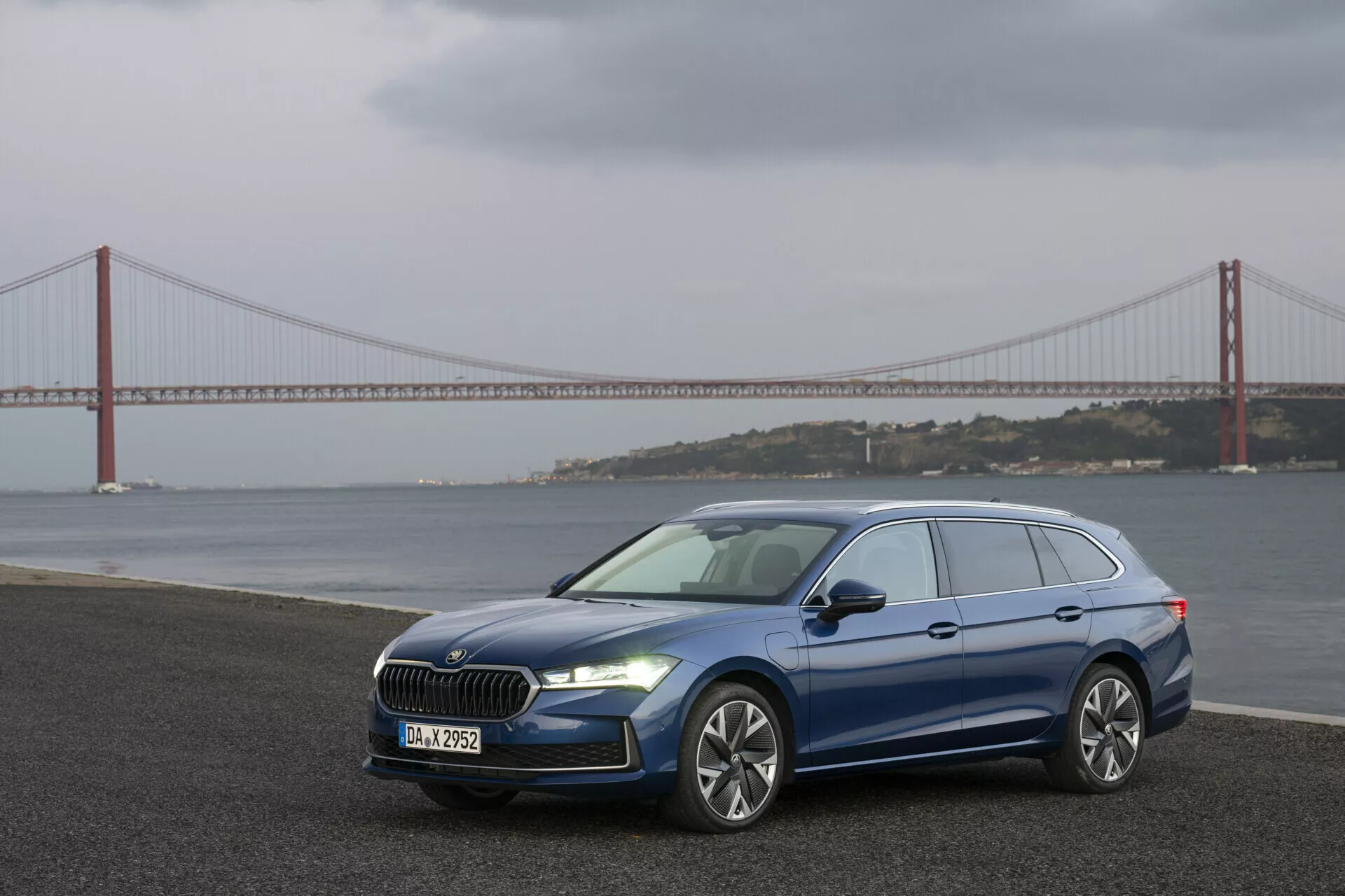 Modrý Škoda Superb Combi 2024 prezentující moderní design s LED světlomety, velkou maskou, aerodynamickým předním nárazníkem s mlhovými světly, slitinovými koly a bočním profilem se střešními nosiči, fotografováno v městském prostředí s pozadím visutého mostu a vody