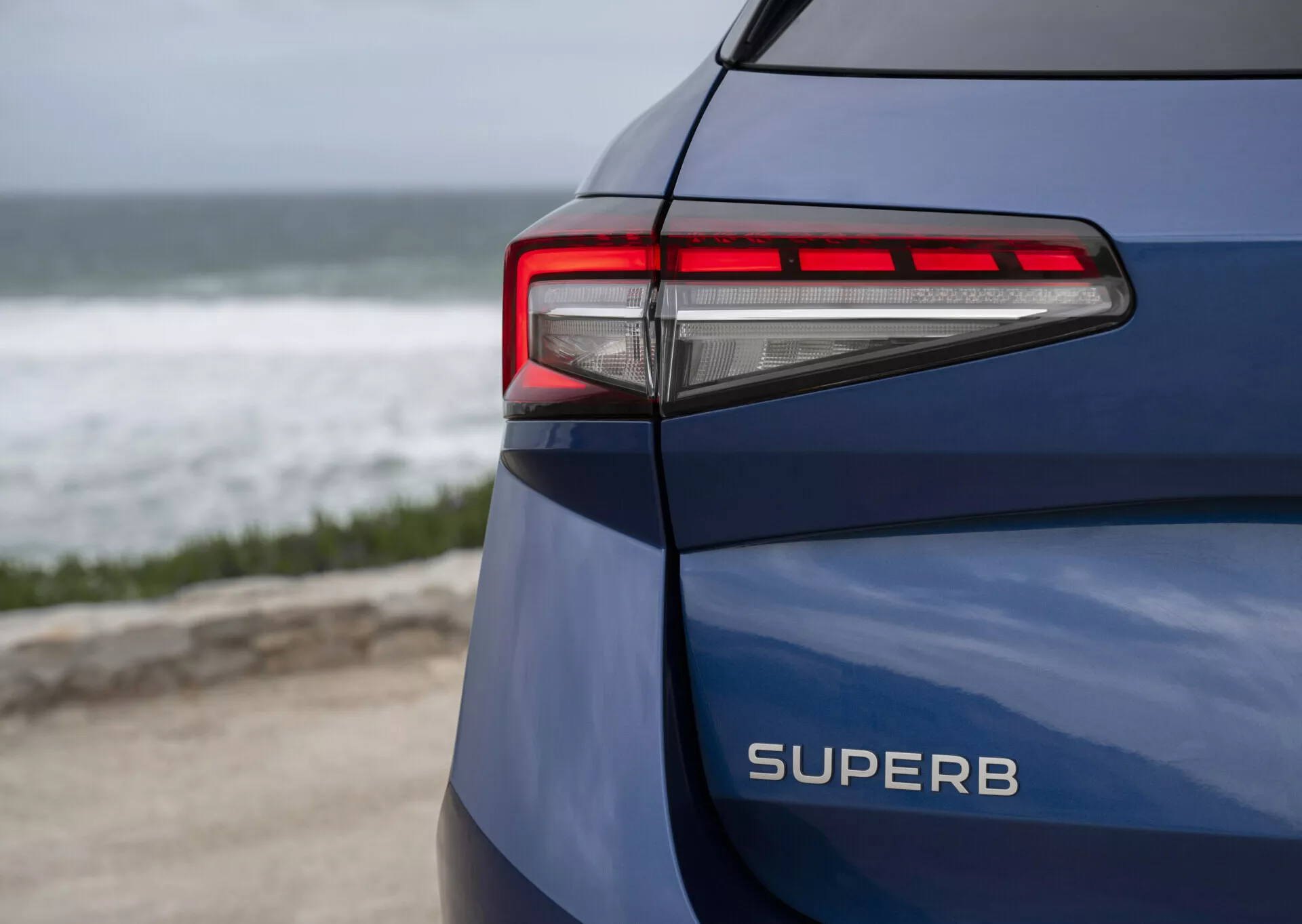 Škoda Superb Combi 2024 v modré barvě, zobrazení zadní levé strany, s jasně viditelným názvem modelu SUPERB, LED zadní svítilna s moderními horizontálními čárami a ostrými úhly na pozadí pobřeží