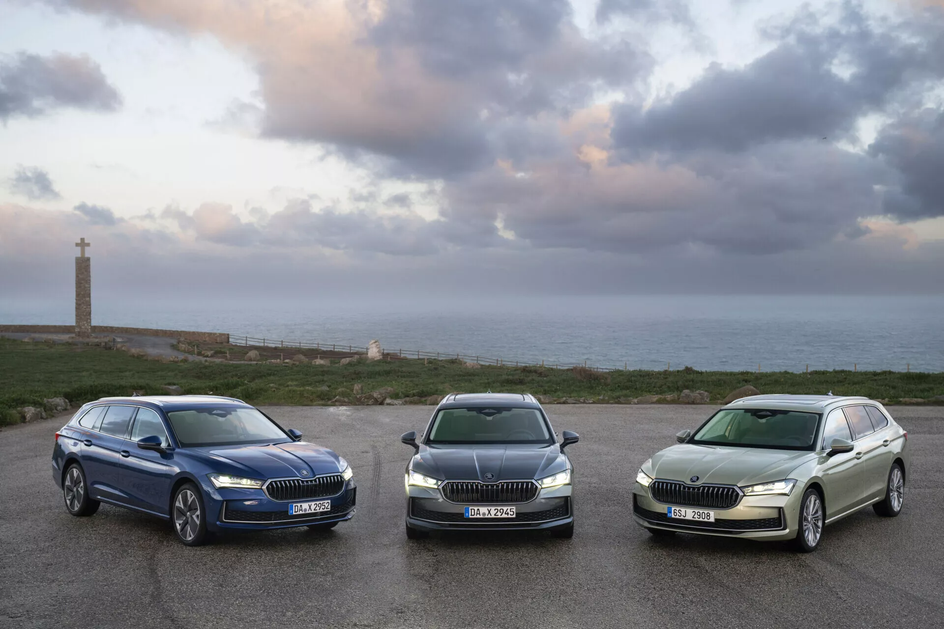 Modely Škoda Superb Combi 2024 zaparkované řadou čelem k foťáku na pozadí pobřežní krajiny, zdůrazňující jejich mřížky, světlomety a boční profily při východu slunce