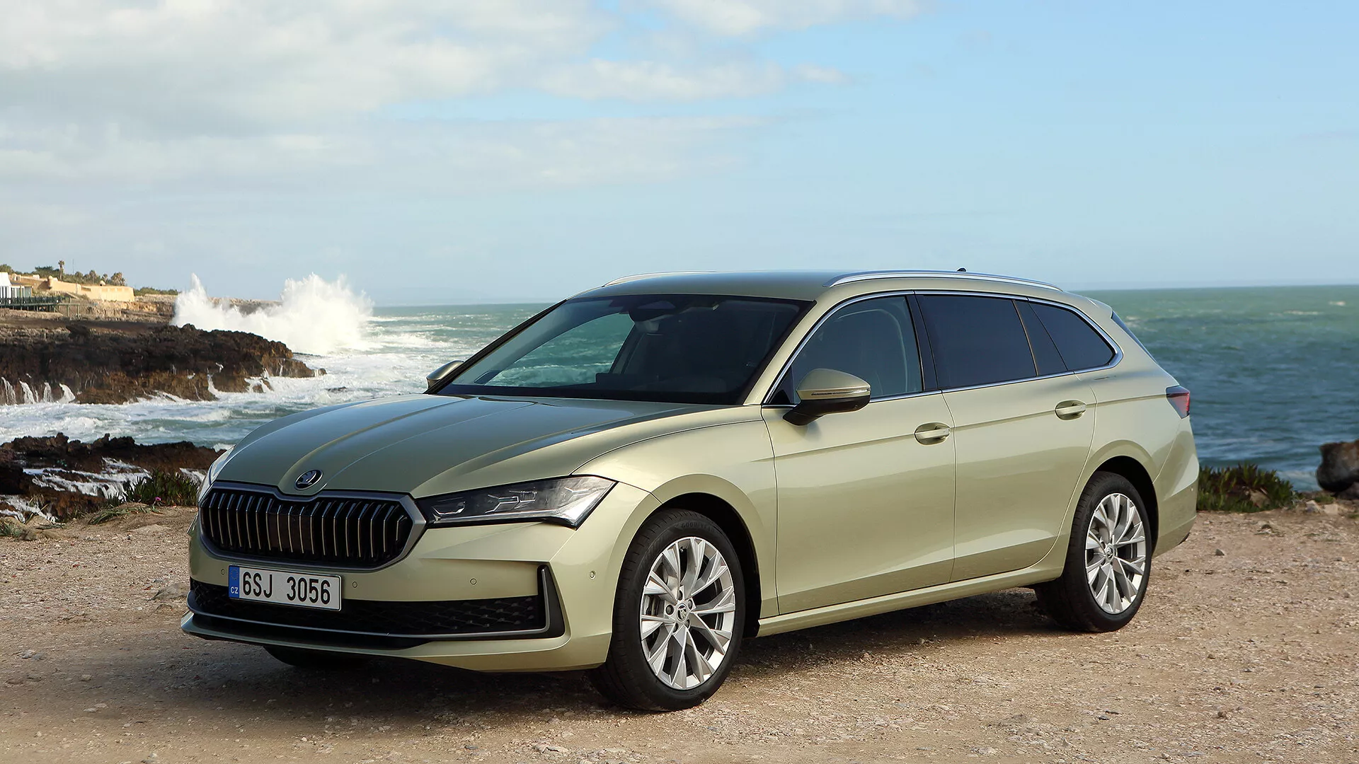 Skoda Superb Combi 2024 představena v přírodním pobřežním prostředí, s výraznou mřížkou chladiče, štíhlými LED světlomety, pokročilými asistenčními systémy pro řidiče a stylovým, avšak praktickým designem s litými koly a střešními nosiči