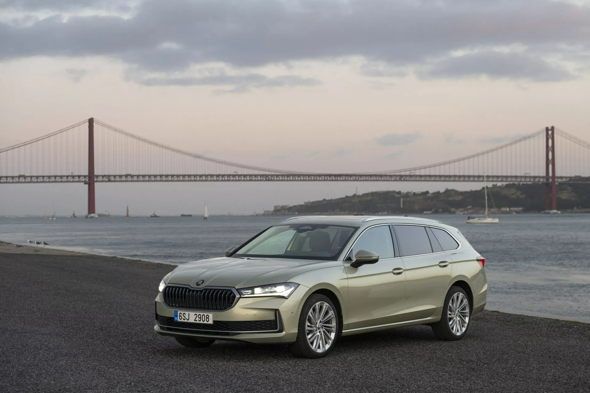 Škoda Superb Combi 2024 předvedená ve tříčtvrtečním předním pohledu zdůrazňující elegantní design hatchbacku, přední mřížku s chromovým povrchem, LED světlomety a slitinová kola na pozadí scenérie s vodní plochou a mostem za soumraku nebo svítání