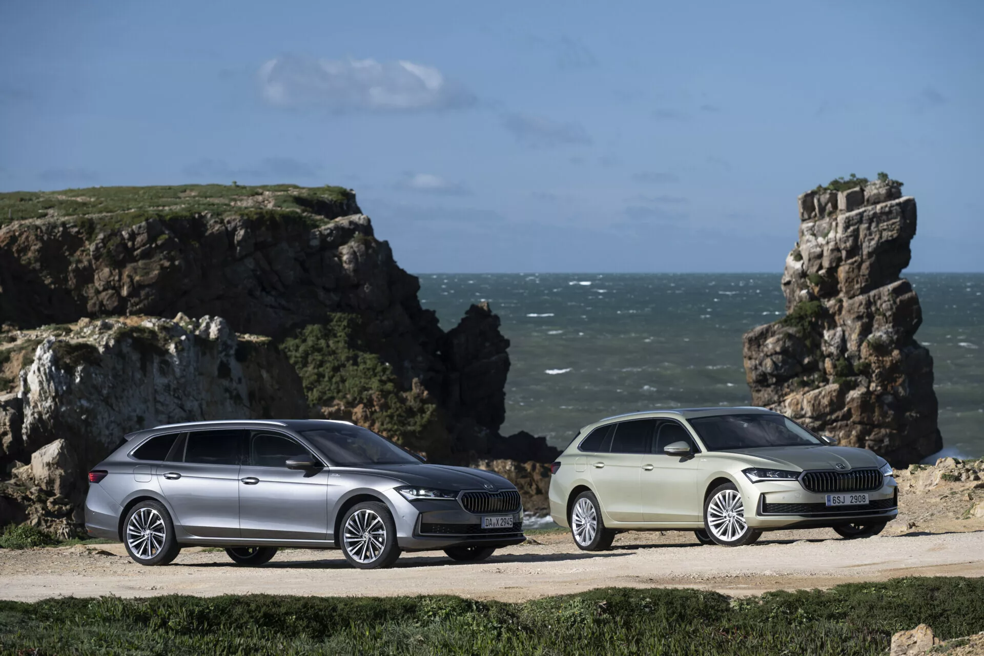 Modely Skoda Superb Combi a sedan roku 2024 zaparkované vedle sebe na pobřežní prašné cestě s pozadím skalnatého mořského útesu a modré oblohy, s prodlouženým prostorem pro zavazadla, aerodynamickým designem, LED světlomety a slitinovými koly, což zdůrazňuje jejich dobrodružný a na životní styl orientovaný přitažlivost.