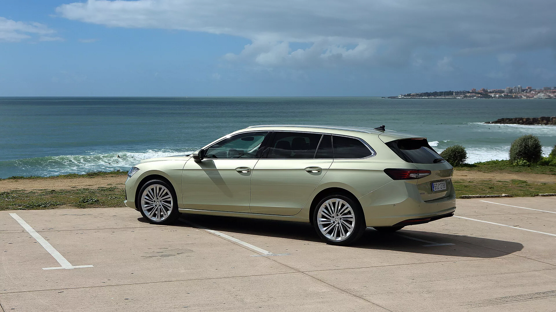 Škoda Superb Combi 2024 v bílé barvě zaparkovaná pod úhlem na parkovišti s výhledem na oceán a pobřeží pod částečně zataženou oblohou, zdůrazňující elegantní design, moderní provedení světlometů a mřížky chladiče, velká slitinová kola a praktické střešní ližiny