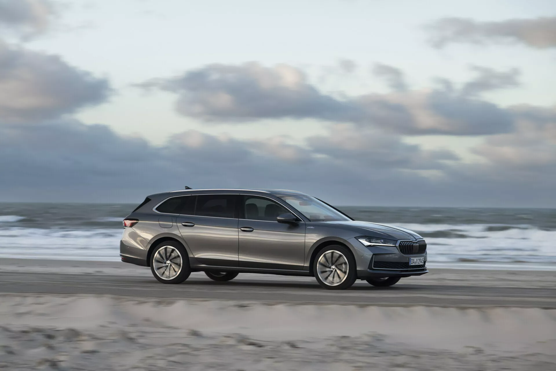 Station wagon Skoda Superb Combi 2024 jedoucí po silnici u pláže, prezentující moderní design s pohledem z boku, rozmazáním pohybu indikujícím rychlost, slitinovými koly a střešními nosiči pro funkčnost.