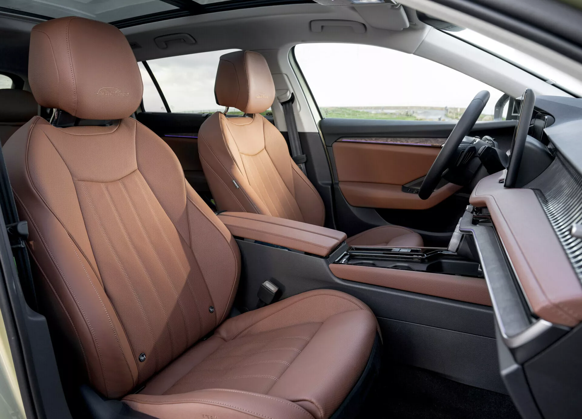 Luxusní sedadla z hnědé kůže a moderní palubní deska v přední kabině Škoda Superb Combi 2024 s automatickou řadící pákou, systémem infotainment a prvky ve dřevěném dekoru z pohledu spolujezdce shora dolů