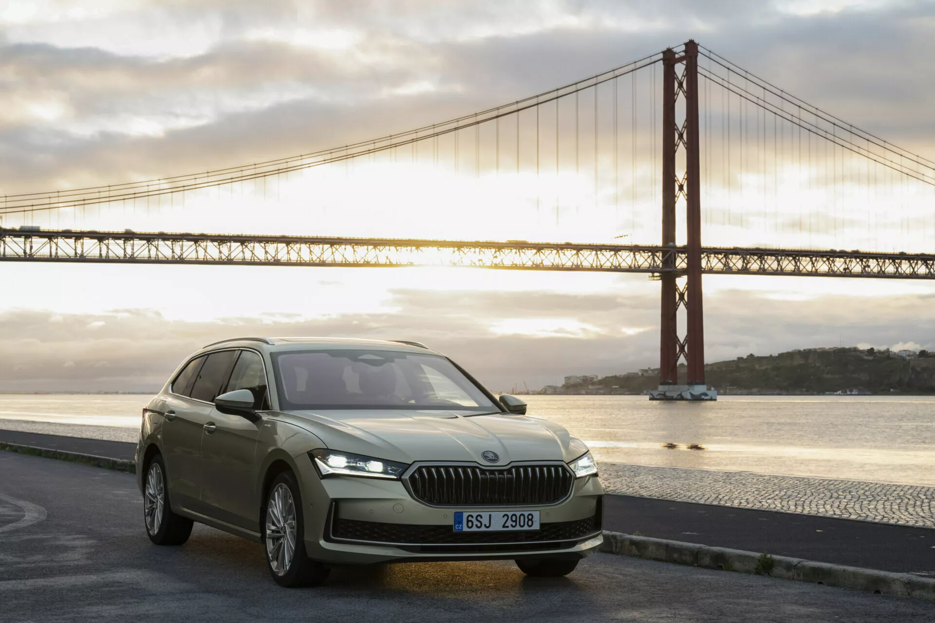 Luxusní SUV Skoda Superb Combi 2024 na svítání s moderními prvkami jako jsou vertikální přední mřížka, štíhlé LED světlomety, tvarovaná kapota a aerodynamický profil u ikonického mostu.