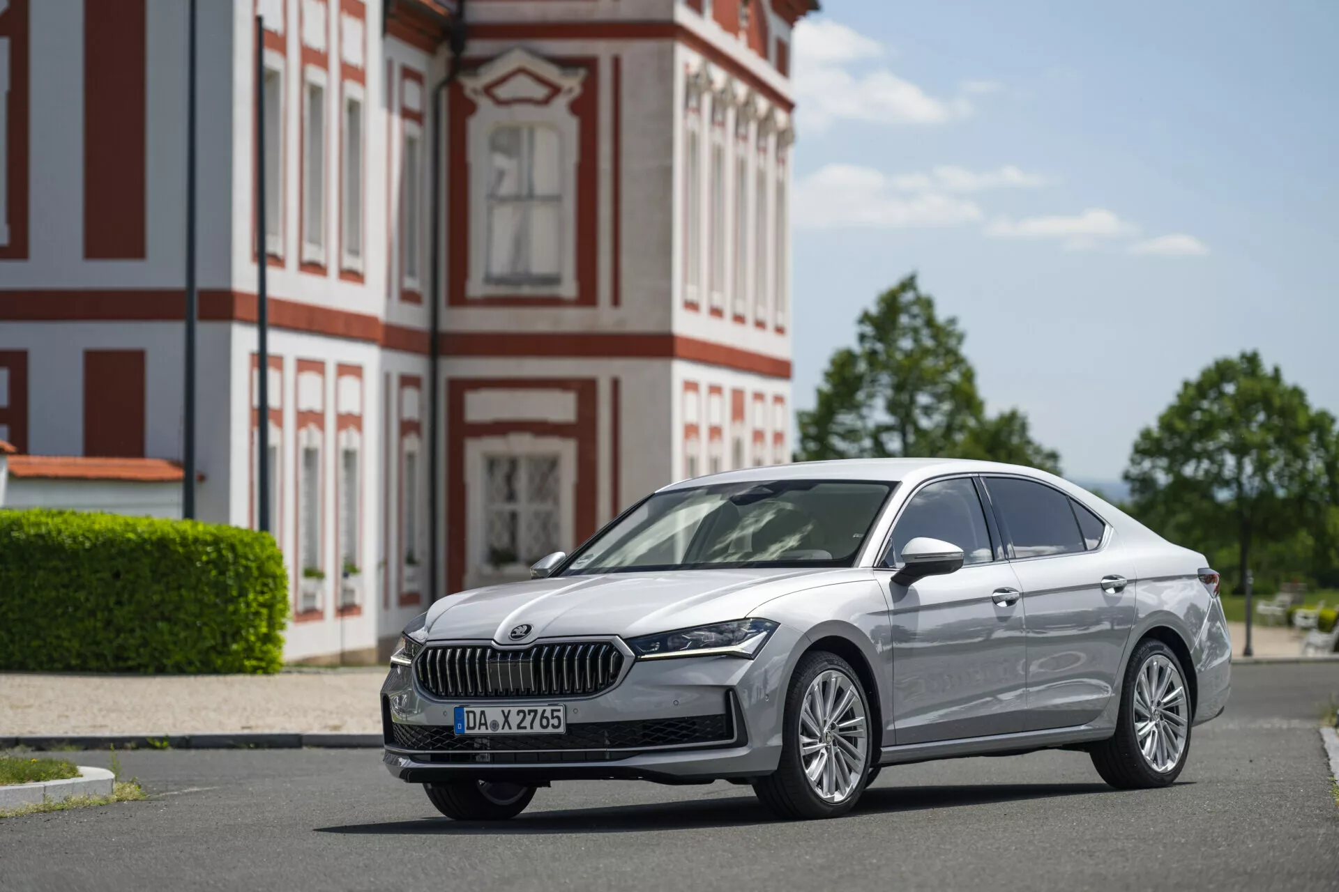 Nízkoúhlý snímek stříbrného sedanu Skoda Superb limuzíny 2024 zaparkovaného na dlážděné silnici. Fotografie zdůrazňuje chromovanou přední masku s vertikálními lamelami, elegantní LED světlomety, mlhovky a vícepaprsková hliníková kola. Boční profil ukazuje čistý design s jemnými linii, chromovými okny a náznaky pokročilého LED osvětlení a bezklíčového vstupu. Historická budova s červeno-bílou fasádou v pozadí přidává na scéně, zatímco čisté modré nebe poskytuje dostatek přirozeného světla k prezentaci funkcí vozu.