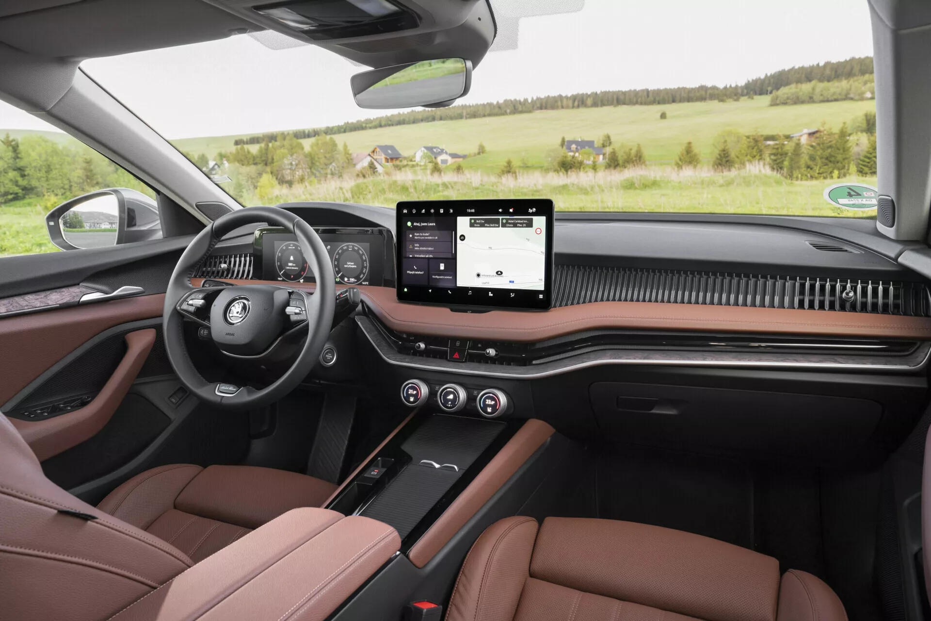 Pohled z první osoby ze zadního sedadla vozu Škoda Superb Limousine 2024, který představuje moderní a luxusní interiér s multifunkčním volantem na levé straně, digitálním přístrojovým panelem, širokým dotykovým infotainment systémem, minimalistickými tlačítky pro ovládání klimatu a sedadel pod ním a karbónovým dekorem. Obrázek také ukazuje dvoubarevná hnědo-černá kožená přední sedadla s kontrastním šitím, odpovídající panely dveří s elektrickými ovládacími prvky, viditelné reproduktory audio systému a panoramatickou střechou, to vše na pozadí venkovské krajiny viděné přes přední čelní sklo.