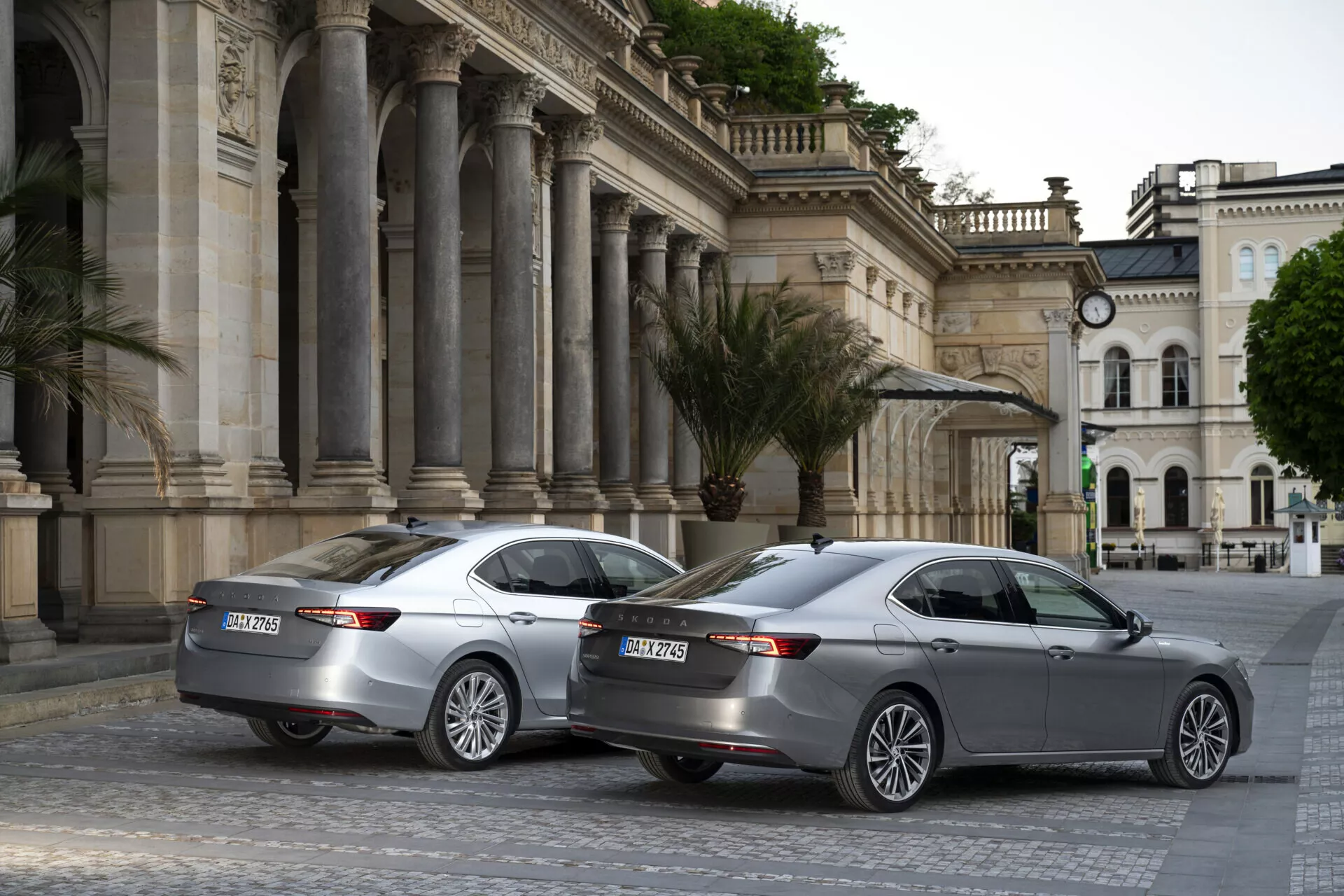 Obrázek zobrazuje dva sedany Skoda Superb 2024 identických modelů zaparkované v urbánním kulturním centru s klasickou architekturou. Snímka pořízená ze tříčtvrtečního zadního úhlu zvýrazňuje jejich elegantní moderní design s LED horizontálními zadními světly, dvojitými výfuky, parkovacími senzory a vícepaprskovými hliníkovými koly. Oba vozy mají žraločí ploutvové antény pro pokročilou konektivitu a třetí brzdové světlo na dolních zadních oknech. Scéna je zachycena za zamračených nebo brzkých ranních/večerních světelných podmínek, což fotografii dodává konzistentní tlumený tón.