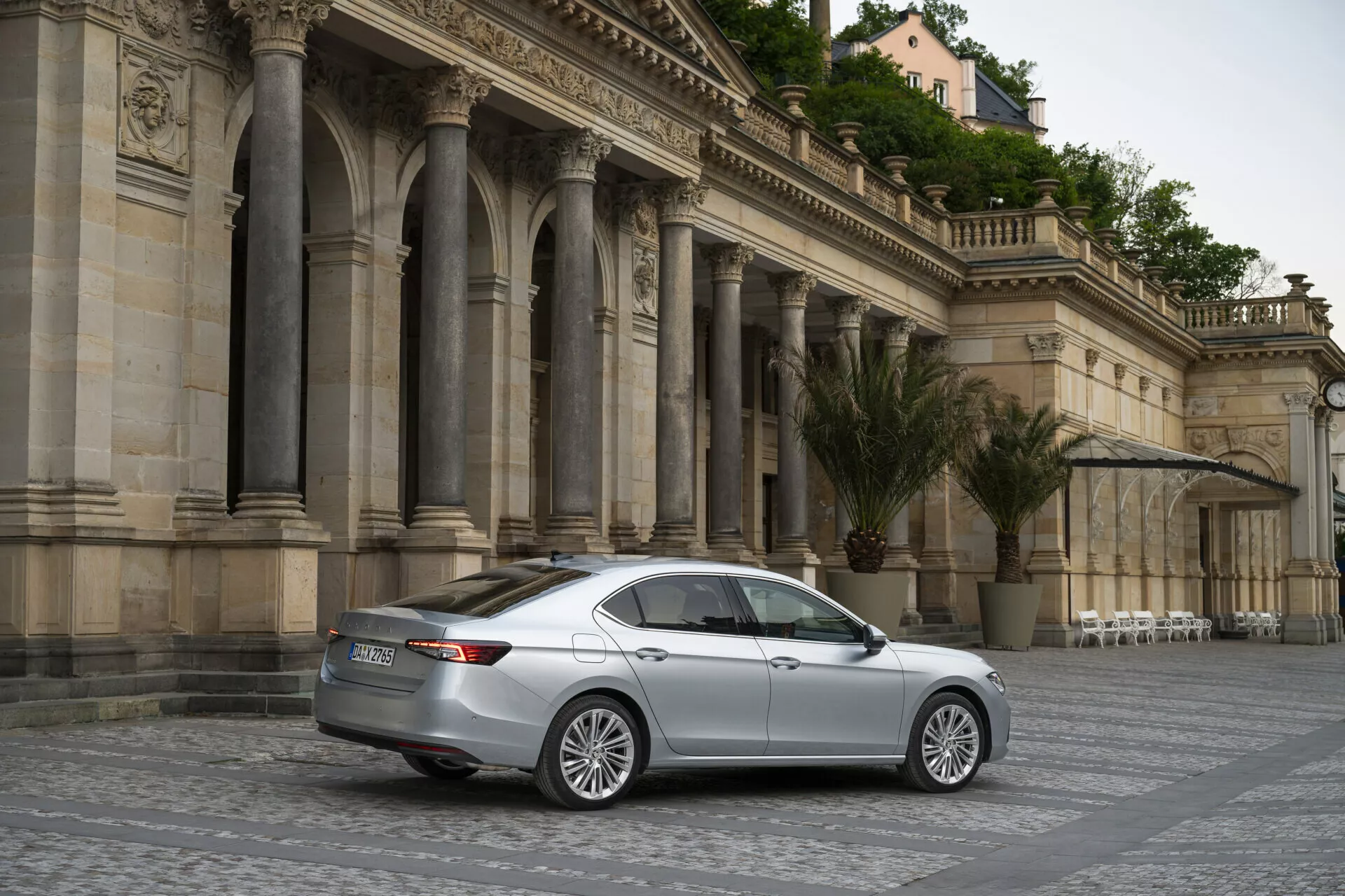Komplexní pohled na stříbrný sedan Skoda Superb Limousine z roku 2024, zaparkovaný diagonálně na dlážděném náměstí s klasickou, zdobenou architekturou v pozadí. Obrázek je pořízen ze zadního tříčtvrtečního úhlu a ukazuje elegantní zadní světla, víko kufru, zadní nárazník a boční kontury vozidla. Měkké osvětlení a klidná atmosféra naznačují zataženou oblohu nebo zlatou hodinu, a stacionární auto s litými koly a designem s více paprsky má kolem sebe dostatek prostoru, což z něj činí vhodný předmět pro kategorie automobilů, luxusních sedanů nebo architektury.