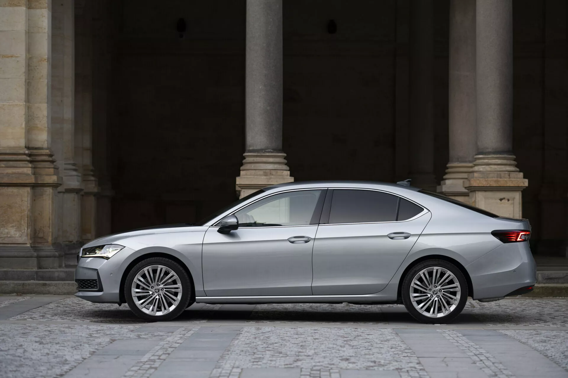 Boční profil vozu Škoda Superb Limousine 2024, čtyřdveřový sedan ve stříbrné/šedé barvě, s hranatými LED světlomety, aerodynamickou přední mřížkou, tónovanými okny, průběžnou linií dveřních klik, vícepaprskovými slitinovými koly a obtočenými zadními světly.