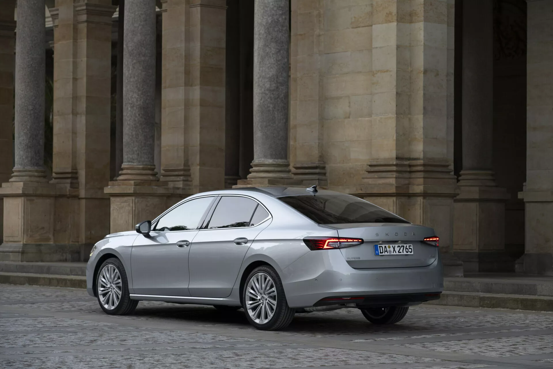 Obrázek sedanu Skoda Superb 2024 z tříčtvrtečního zadního úhlu. Zvýrazňuje pravou stranu a zadní část, vůz má hliníková kola, LED zadní světla, aerodynamicky optimalizované tělo s metalickou barvou, tónovaná okna a viditelný znak. Obrázek, pořízený ze střední vzdálenosti, ukazuje vozidlo bez překážek s klasickou architekturou v pozadí.