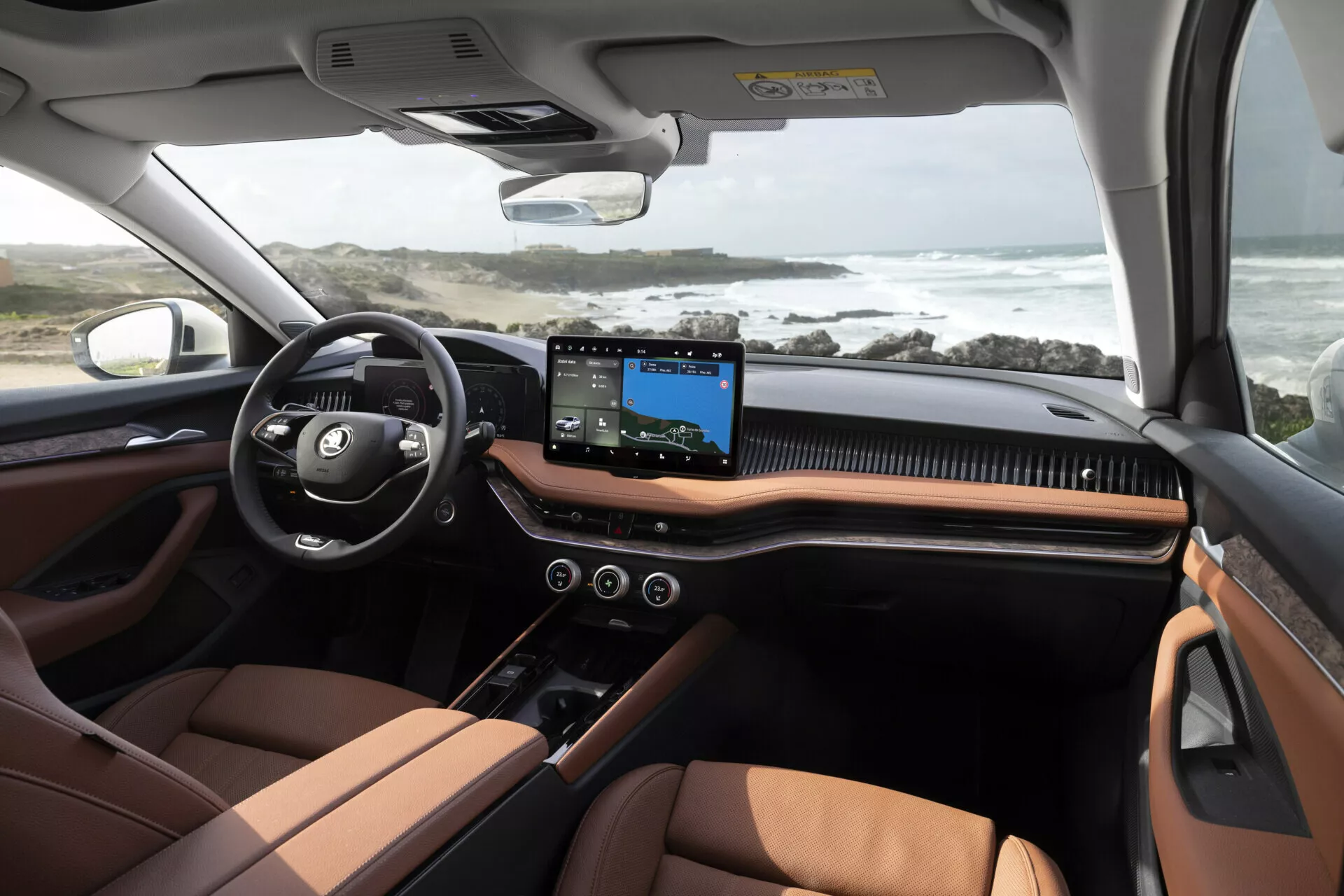 Pohled do interiéru modelu Škoda Superb Limousine 2024 z prostoru zadních sedaček, prezentující luxusní a moderní kabinu. Palubní deska obsahuje velký centrální dotykový displej s navigační mapou, multifunkční volant a dvoubarevné schéma černé a bohatě hnědé, pravděpodobně v koženém provedení. Technologické prvky zahrnují ovládání klimatizace, bezrámové zpětné zrcátko a ovládání panoramatického střešního okna. Pobřežní scenérie viditelná z oken dodává snímku životní styl.
