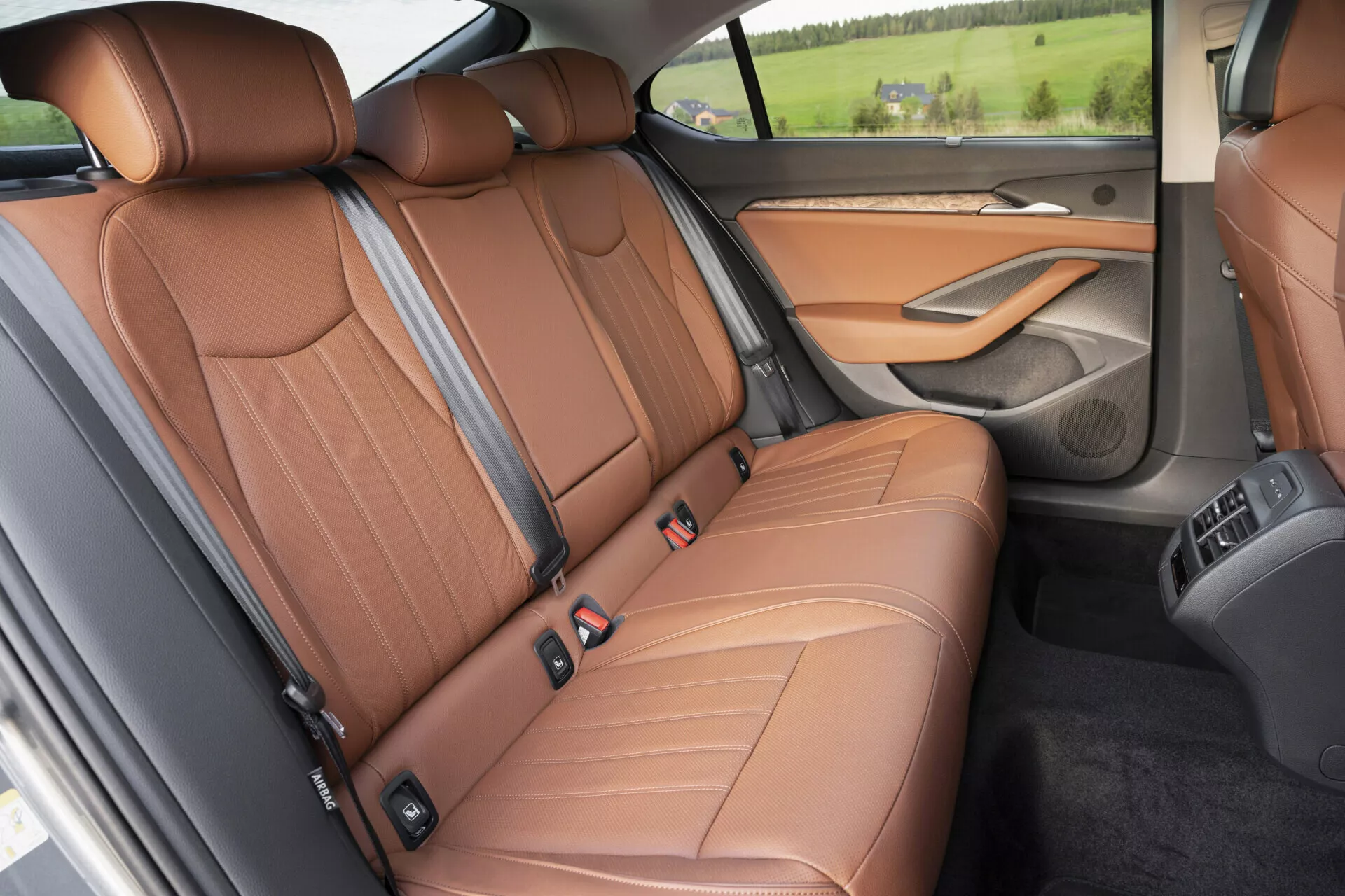 Detailní pohled na zadní prostor pro cestující modelu Skoda Superb 2024 se zaměřením na luxusní zadní sedadlo z hnědé kůže s výrazným prošíváním, opěrkami hlavy pro tři cestující, skládací středovou opěrkou, viditelnými ovládacími prvky klimatizace a dřevěným dekorem dveřního panelu, zachycený z pohledu shora a lehce zboku.