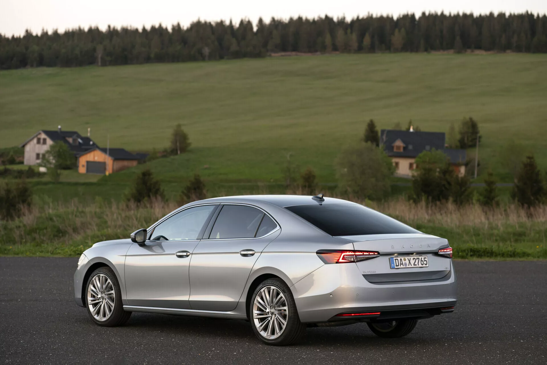 Obrázek zobrazuje 2024 Skoda Superb Limousine, moderní sedan s elegantním, aerodynamickým designem a kovovým stříbrným povrchem, zachyceným z zadního tříčtvrtečního úhlu. Auto stojí na hladké asfaltové silnici v venkovském prostředí s otevřenými poli a domy v pozadí. Významné rysy zahrnují obklopující LED zadní světla, zadní spoiler, vícepaprsková litá kola, odmlžovací mřížku na zadním skle a žraločí ploutev na střeše. Fotografie, pořízená během zlaté hodiny, má mělkou hloubku ostrosti zaměřenou na auto, přičemž přirozené světlo zvýrazňuje jeho obrysy.