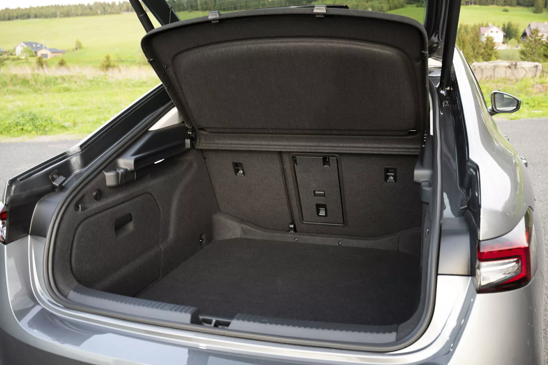 Detailní pohled na otevřený kufr vozu Skoda Superb 2024 sedan, ukazující prostornou, kobercem pokrytou nákladovou oblast s dělenými zadními sedadly, háčky na zavazadla, osvětlením kufru, přístupovými panely, zásuvkou a vysouvacím krytem na ochranu soukromí, na pozadí přírodního venkovního prostředí.