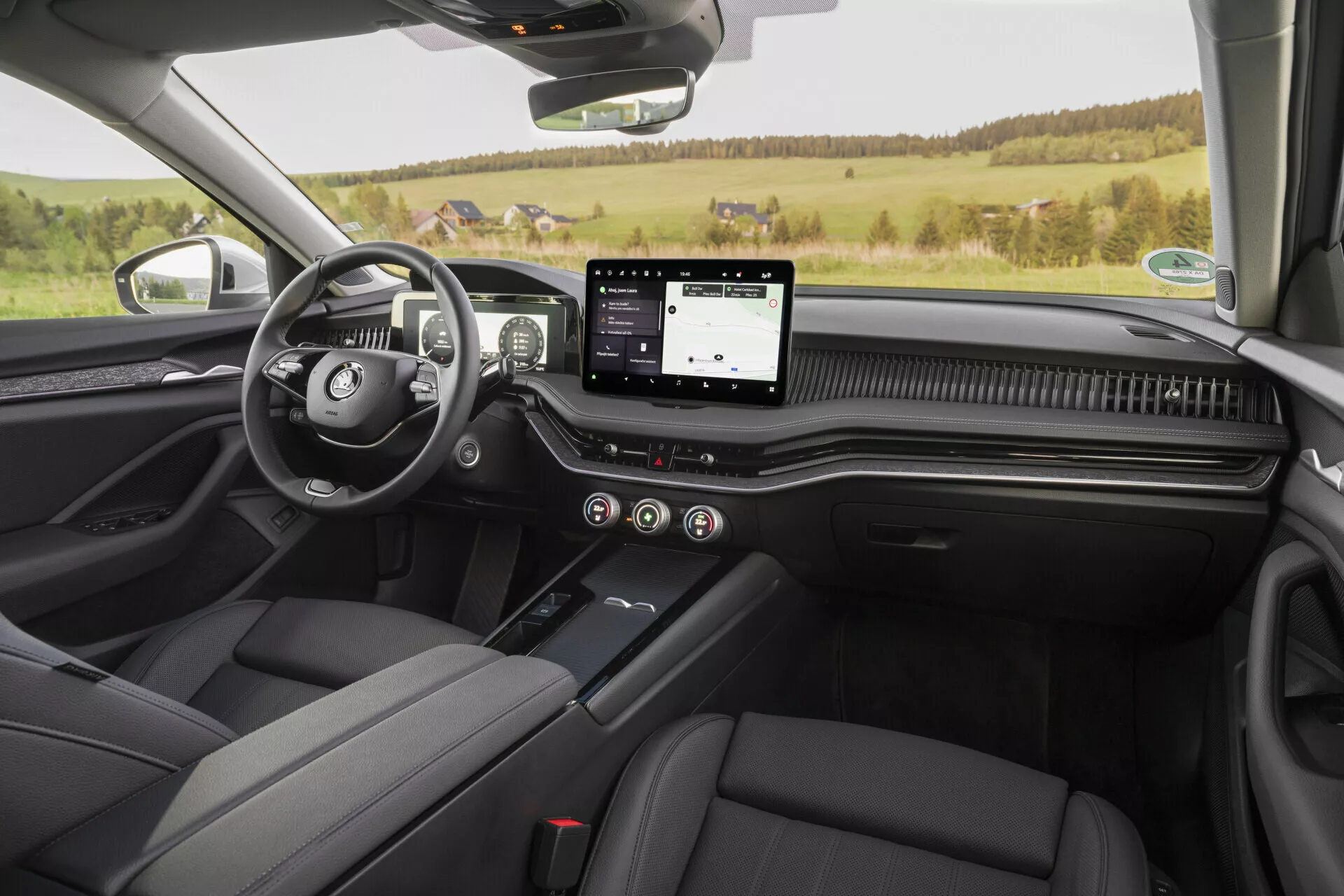 Interiér vozu Skoda Superb 2024 limuzína z pohledu zadního sedadla, zobrazující přední palubní desku, sedadla řidiče a spolujezdce v černé kůži, centrální konzolu s řadicí pákou, dotykový infotainment systém, plně digitální přístrojovou desku, karbonové akcenty a klidné venkovské prostředí venku.