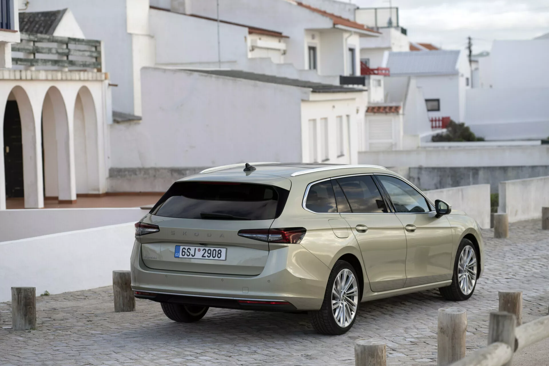 Obrázek ukazuje elegantní kombi Škoda Superb 2024 z třičtvrtinové zadní perspektivy, zaparkované na dlážděné dlažbě s několika krátkými sloupky působícími jako zábrany. Detaily zahrnují moderní LED zadní světla, prostorné zadní sklo s stěračem, parkovací senzory v zadním nárazníku, osobitá litá kola, stříbrné střešní nosníky a 'žraločí ploutev' anténu. Boční profil odhaluje prodlouženou zadní část, indikující dostatek prostoru pro náklad. Přirozené denní světlo zvýrazňuje design a užitnost vozidla v možném prostředí malého města nebo vesnice.