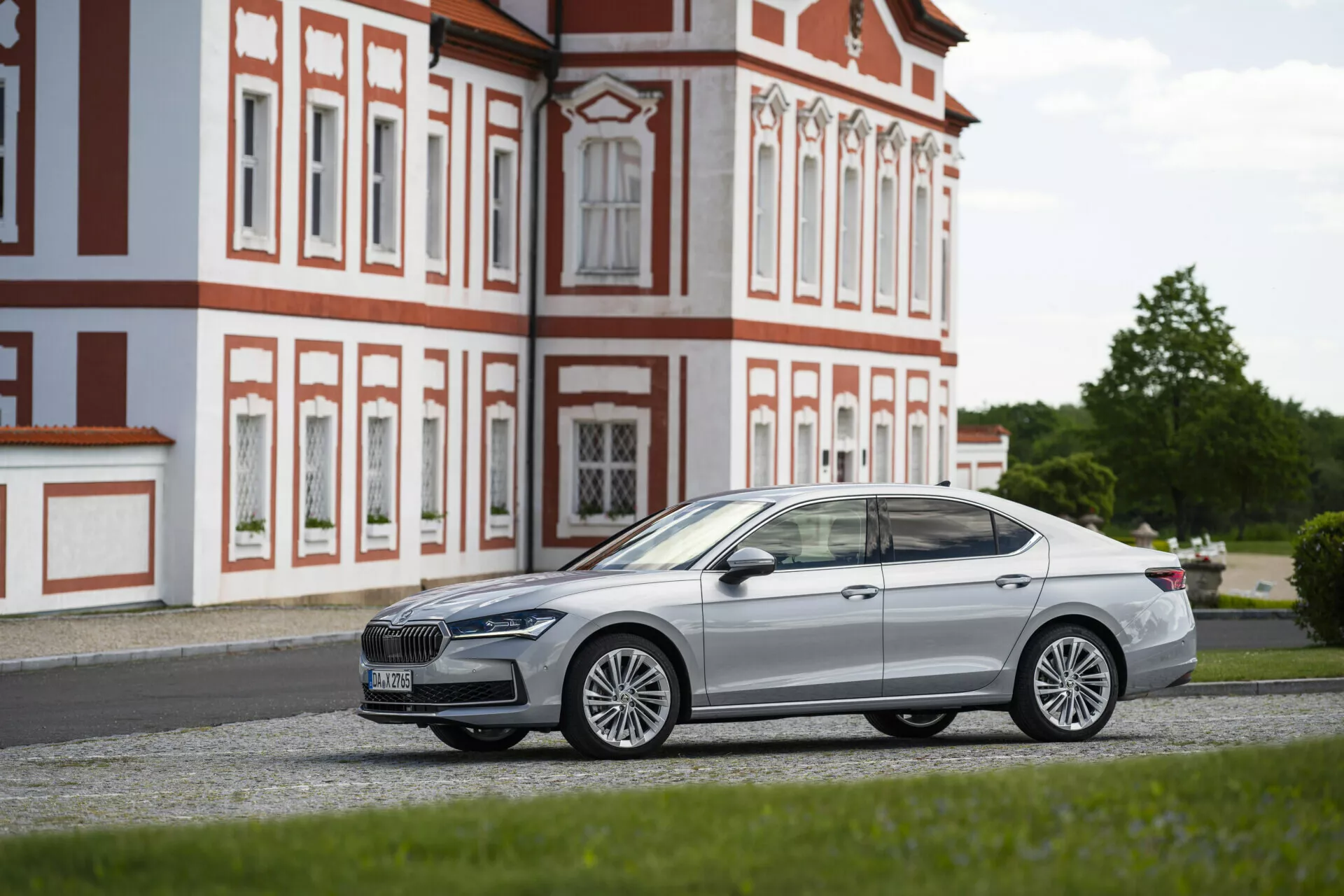 Boční profil moderního sedanu zaparkovaného na štěrku, zaměřený na stranu spolujezdce, pořízený z nízkého úhlu. Auto, umístěné paralelně s kamerou, předvádí svůj elegantní design, chromované akcenty, stylová litá kola a výraznou přední masku s viditelným logem. Hladká, aerodynamická silueta s definovanými liniemi, sklopenými zpětnými zrcátky a zapadnutými klikami dveří naznačuje pokročilý bezklíčový vstup. Zatmavená okna naznačují tónovaná skla nebo UV ochranu. Barokní budova v pozadí kontrastuje s moderností vozu.