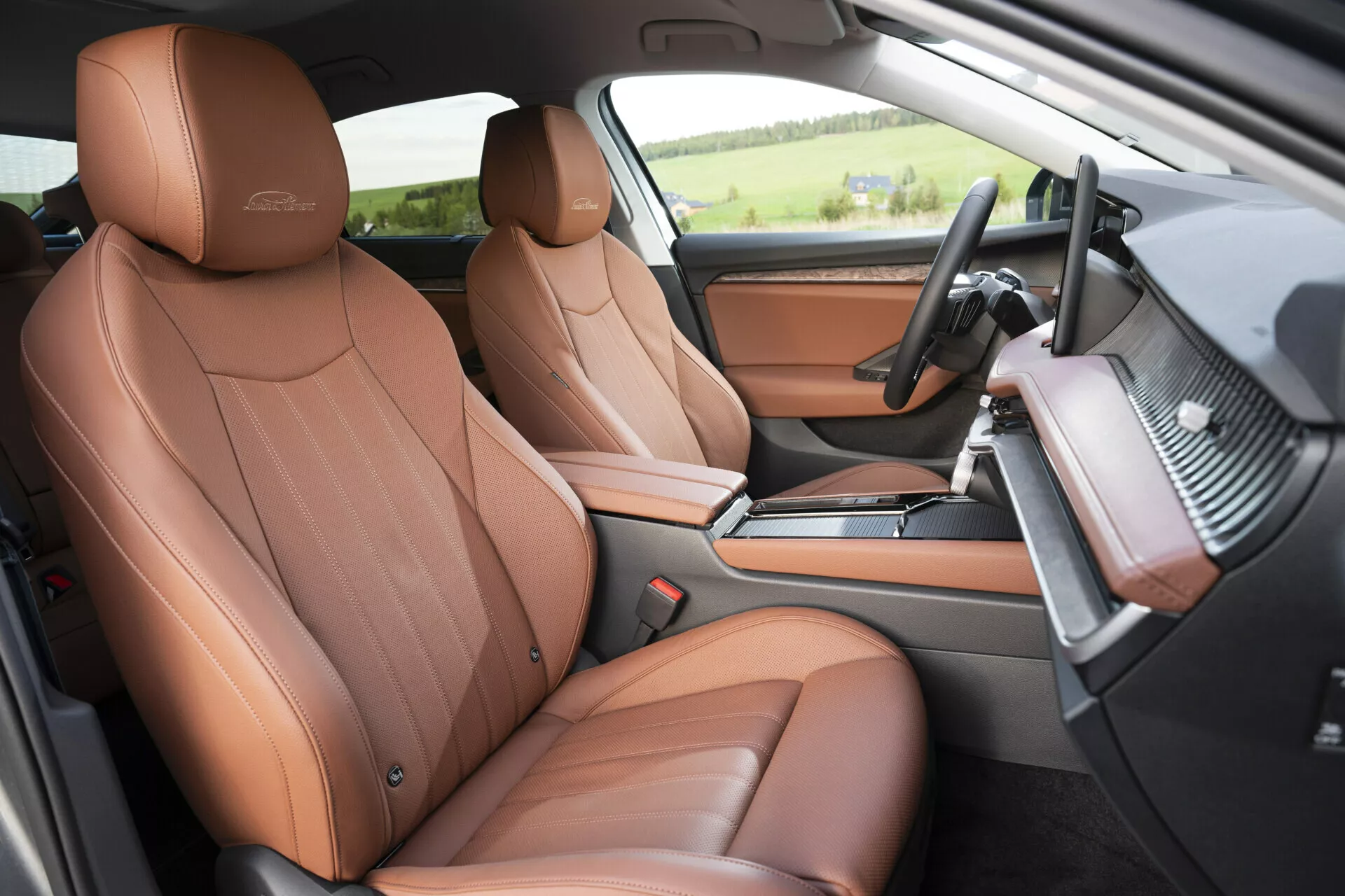 Tento obrázek zobrazuje interiér vozu Skoda Superb Limousine 2024, zaměřující se na přední část pro řidiče a spolujezdce, pohledem z mírně zvednuté perspektivy od spolujezdce. Ukazuje prémiová hnědá kožená sedadla s luxusním embosováním, multifunkční volant potažený kůží s ovládacími tlačítky a plochým spodním designem, přístrojovou desku s uhlíkovým vláknem a možným ambientním osvětlením a středovou konzolu s řadicí pákou, držáky na nápoje a úložným prostorem. Vnitřek dveří odpovídá hnědé kůži sedadel s kartáčovaným kovovým zdobením. Podlaha je kobercovaná pro větší pohodlí a lepší zvukovou izolaci, což zdůrazňuje luxusní a technologicky vyspělý interiér vozu.