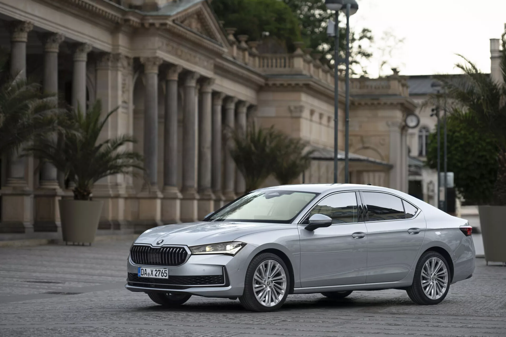 Tento obrázek zobrazuje moderní sedan Skoda Superb 2024 zaparkovaný na vydlážděné otevřené ploše, pravděpodobně na náměstí nebo nádvoří, s klasicky navrženou budovou s pilíři v pozadí, evokující formální prostředí, možná vládní budovy nebo kulturní místa. Auto, snímáno z tříčtvrtečního předního úhlu, lehce nakloněné směrem k řidičově straně, nabízí jasný pohled na přední a boční profil, zdůrazňující desénové prvky jako výrazná mřížka s konkrétním vzorem, elegantní horizontální LED světlomety, mnohopaprsková litá kola, aerodynamické linie karoserie, zpětná zrcátka s integrovanými směrovými světly a metalický lak. Emblém značky na mřížce a možná zadní specifické nápisy podtrhují prémiové postavení vozu.