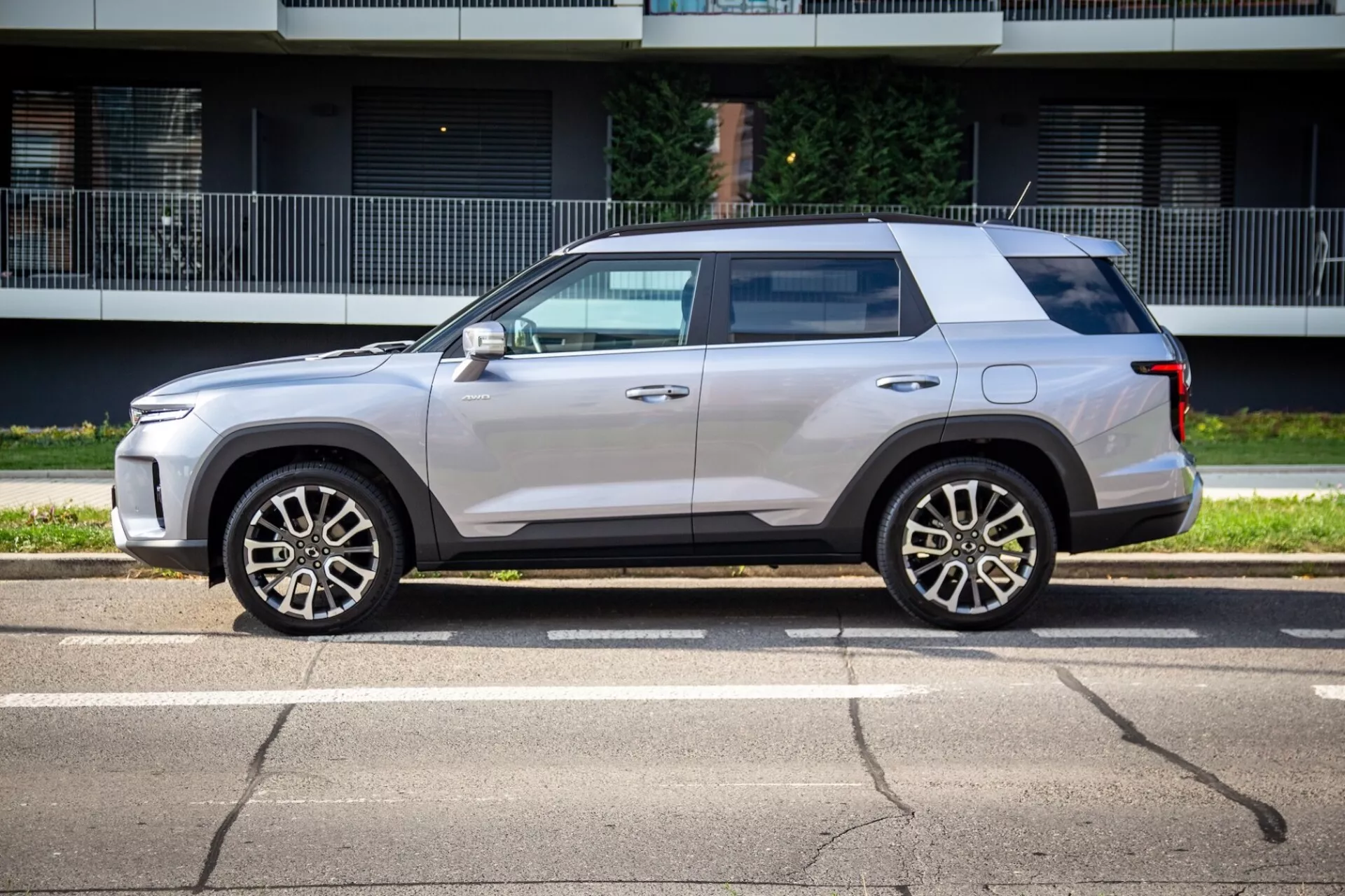 Profilový pohled na SsangYong Torres 2023 ukazující jeho hranatý, přesto aerodynamický design, vysokou linii pasu, plochou střechu a světle metalický lak s pěticípkovými slitinovými koly, zrcátka v barvě karoserie s integrovanými směrovými signály, mírné tónování oken, zapuštěné kličky dveří a černé obložení na slunné ulici
