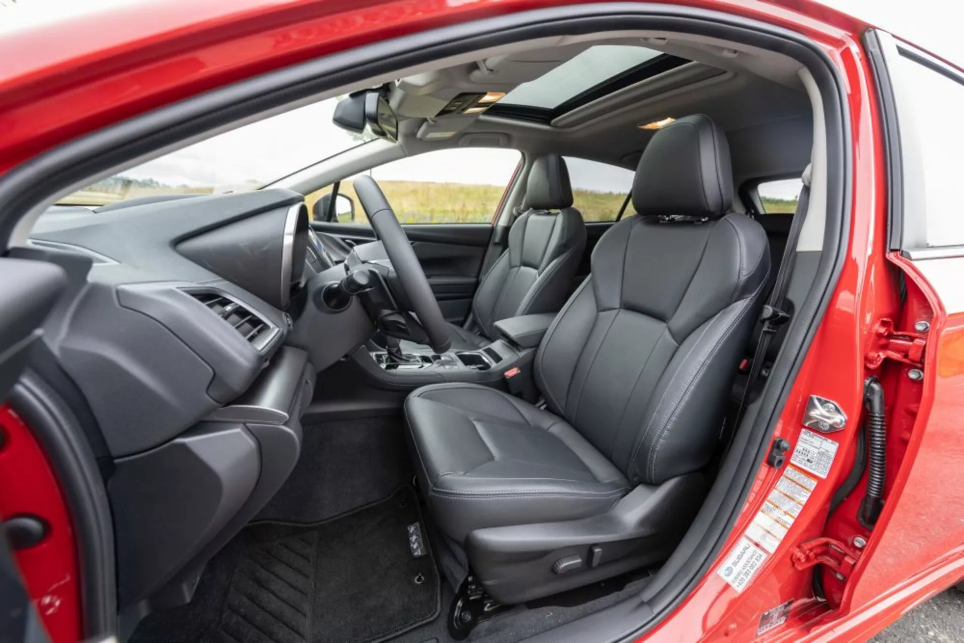 Interiér Subaru Impreza 2018 z pohledu z venkovní strany ukazuje sedadlo řidiče, palubní desku, volant s ovládacími tlačítky, čalounění předních sedadel v černé kůži s ergonomickým designem a ovládání na vnitřní straně dveří skrze otevřené dveře řidiče
