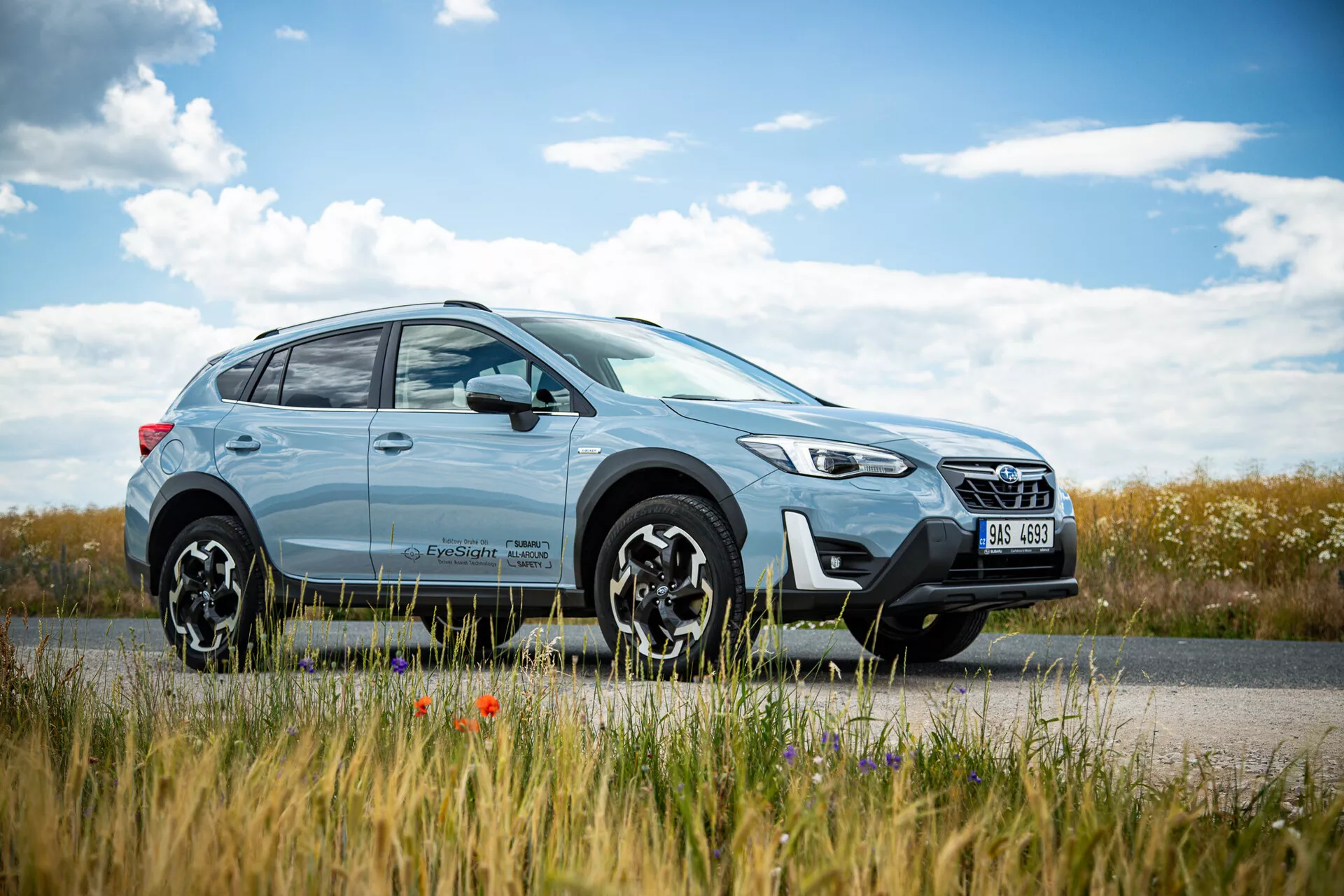 Světle modrý křižník SUV Subaru XV 2021 zaparkovaný u silnice s krajinou venkova na pozadí, s moderním designem s LED světlomety a slitinovými koly s více paprsky, zdůrazňující schopnosti jízdy ve městě i v terénu.