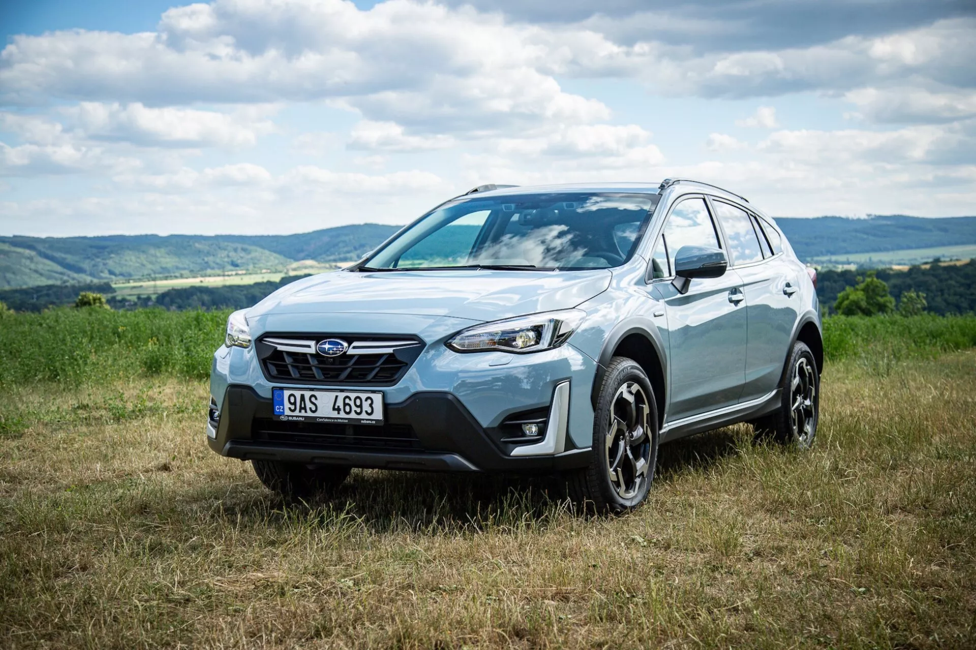 Crossover SUV Subaru XV 2021 zaparkovaný na trávě s kopcovitým pozadím, zdůrazňující přední masku, profil strany a design kola