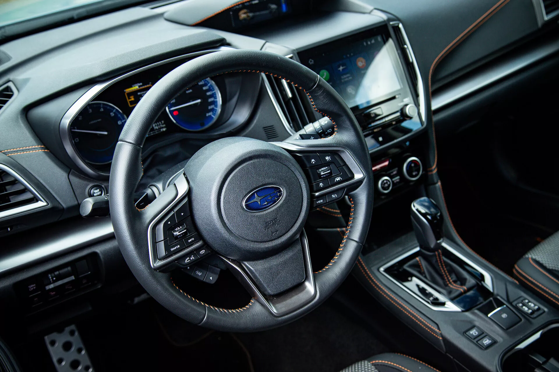 Interiér Subaru XV 2021 z pohledu předního spolujezdce, zobrazuje tříramenný volant s multifunkčními ovladači, přístrojový štít s analogovými a digitálními displeji na palubní desce, centrální dotykový systém infotainment, tlačítka klimatizace, páku automatické převodovky a luxusní materiály, včetně kůže a chromových akcentů.