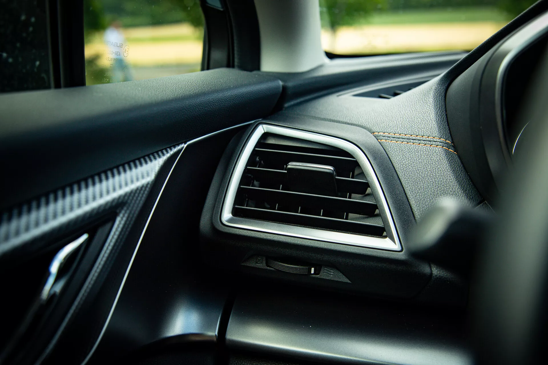 Detail interiéru Subaru XV 2021 zobrazující palubní desku na straně spolujezdce s chromově zdůrazněným větracím otvorem, kvalitním černým kůženým čalouněním se zřetelným prošíváním a materiály soft-touch, fotografovaný pod úhlem 45 stupňů, který zdůrazňuje moderní designové prvky ve dnešním denním světle