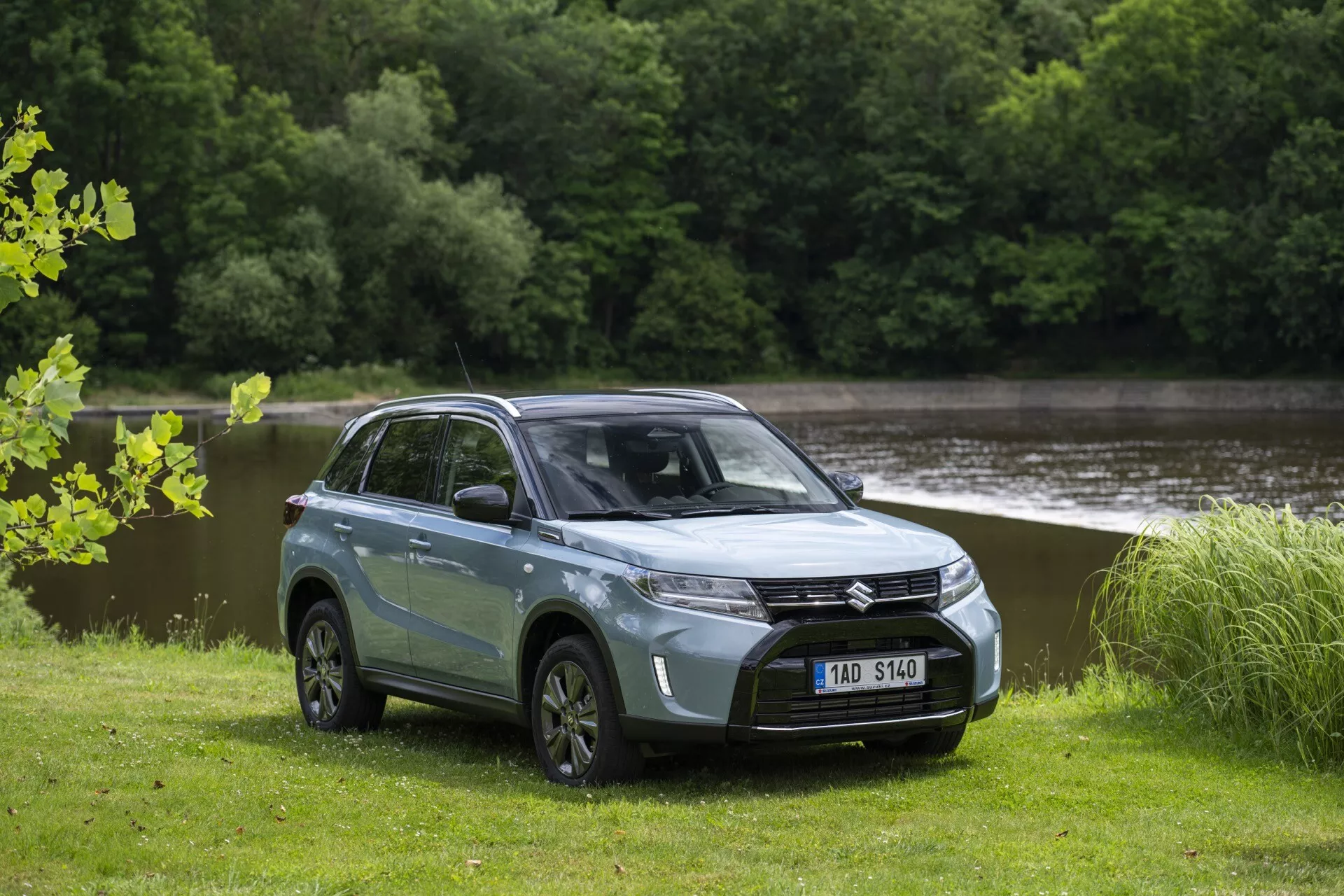 Fotografie moderního SUV Suzuki Vitara 2024 (2. generace, facelift 2) v dvoubarevném laku, svítivě modré s černou střechou. Zaparkované na trávě s vodou a stromy v pozadí. SUV je zobrazeno z čelního kvadratového úhlu, ukazuje řidičovu stranu. Má výraznou přední masku s centrálním emblémem, elegantní úhlová LED světla, hladkou kapotu, vícepaprsková kola z lehkých slitin a viditelnou světlou výšku. Tónovaná okna, boční zrcátka a barevně sladěné kliky dveří podtrhují moderní design, zatímco zrcátka mají integrované LED směrovky.