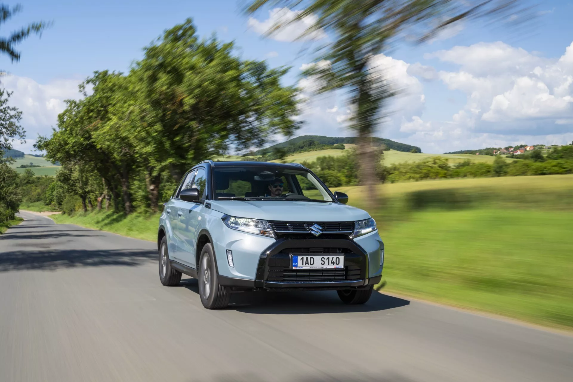 Suzuki Vitara 2024 2. generace Facelift je zachycen v pohybu s rozmazaným pozadím stromů a oblohy, což zdůrazňuje jeho rychlost. Tento pohled z předního šikmého úhlu zvýrazňuje moderní SUV se zvýšenou světlou výškou, signaturní mřížkou, LED denními světly, mlhovými světly a vzduchovými otvory. Boční zrcátka, aerodynamické tělo a pohybující se litá kola zdůrazňují jeho design. Viditelné senzory naznačují pokročilé asistenční systémy řízení, jako je adaptivní tempomat a vyhýbání se kolizím. Kovový lak, čistý vzhled a jasná, ale anonymní poznávací značka přispívají k jeho estetice. Pozice na venkovské silnici ukazuje všestrannost vozidla pro různé druhy jízd.