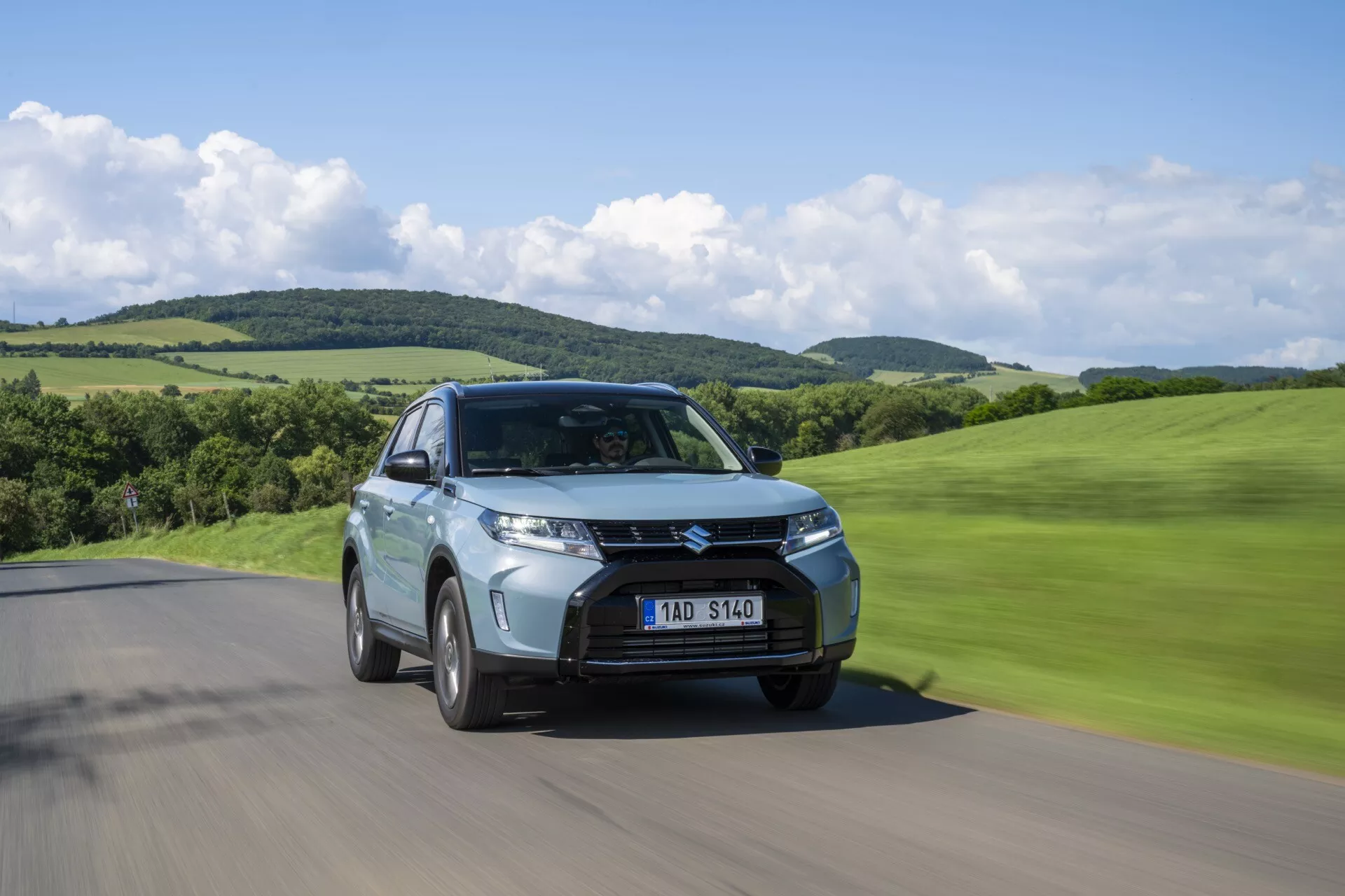 Nízký úhel záběru Suzuki Vitara 2024 druhé generace facelift na silnici, přední tříčtvrtinový pohled zobrazující mřížku, světlomety a pohybovou neostrost v zelené, kopcovité krajině.