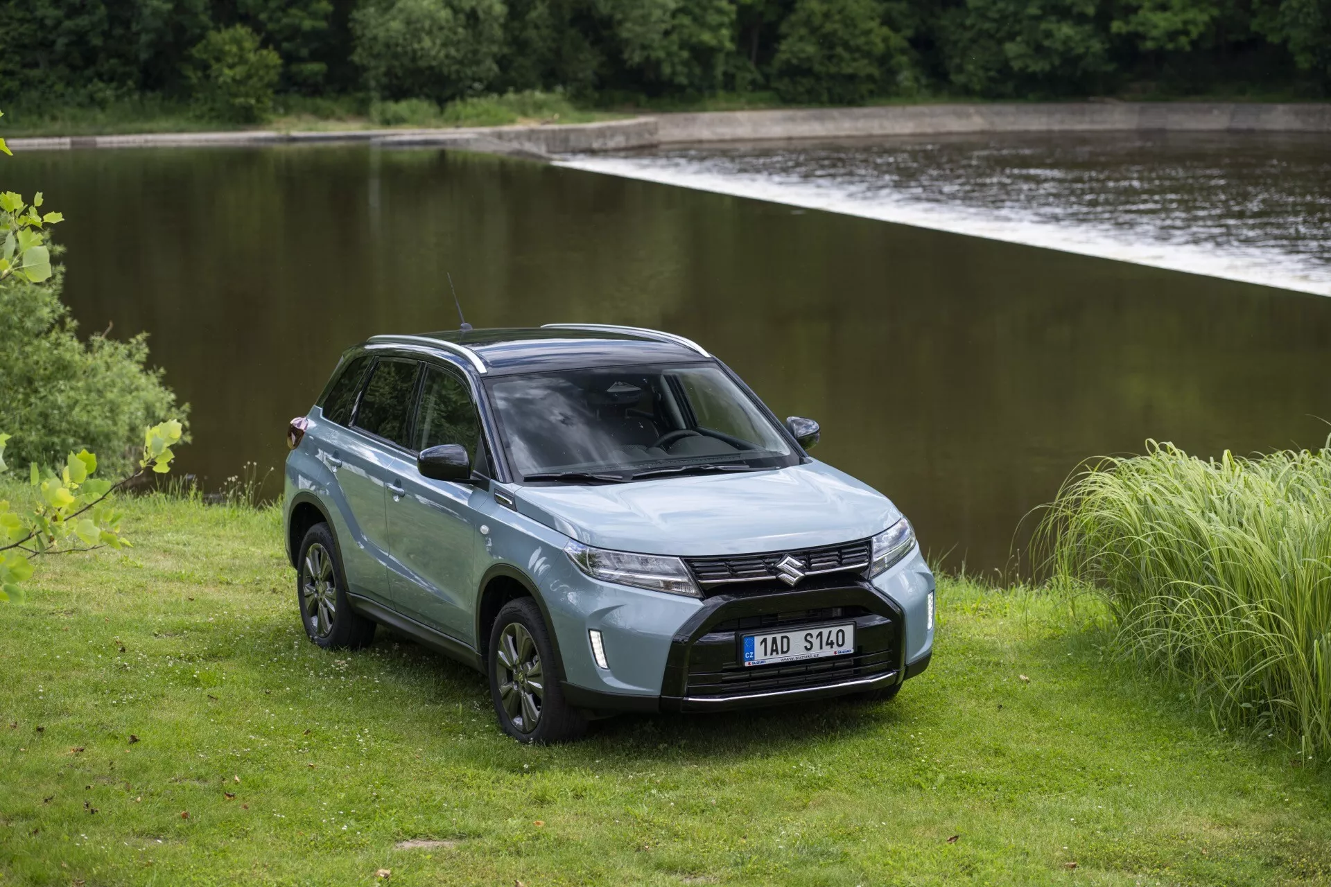 Na obrázku je SUV Suzuki Vitara 2024 (druhá generace, druhý facelift) v klidném venkovním prostředí u vodní plochy. Pořízený ze tříčtvrtečního předního pohledu, SUV má viditelná moderní LED světla, mřížku chladiče a přední nárazník. Sochařsky vykrojená kapota a skloněné čelní sklo přidávají aerodynamický profil. Na jedné straně vozidla je vidět čtyřdveřový design, zavřený nákladní prostor, litá kola a robustní terénní pneumatiky. Tlumená stříbrná nebo šedá barva dodává eleganci. Nezpevněný přírodní terén kolem SUV podtrhuje jeho schopnosti mimo silnici a dobrodružnou povahu.