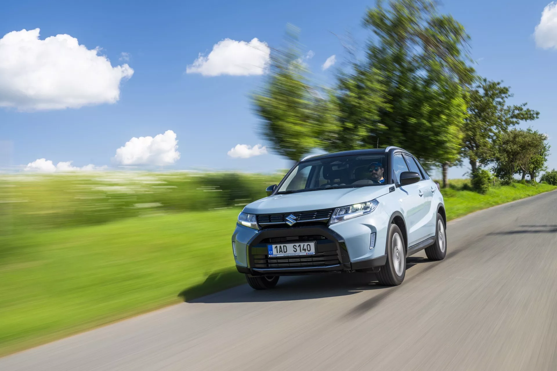 Dynamický záběr Suzuki Vitara 2024 (2. generace, facelift) zachycený z předního tříčtvrtečního pohledu. Auto jede po silnici s rozmazanou zelenou krajinou v pozadí, což naznačuje pohyb. Viditelné prvky zahrnují kapotu, mřížku chladiče, světlomety, přední nárazník a mírně natočené přední kolo. Řidičova strana, včetně dveří a bočního zrcátka, je výrazně vidět. Pohybové rozmazání a pruhy podél okolí zvyšují pocit rychlosti, zatímco auto zůstává zaostřeno. Patrné jsou kontury, řezy na straně a design kol. Jasné denní světlo s čistou oblohou a stínem naznačuje sluneční světlo z levého horního směru. Přes čelní sklo lze vidět postavu, pravděpodobně řidiče.