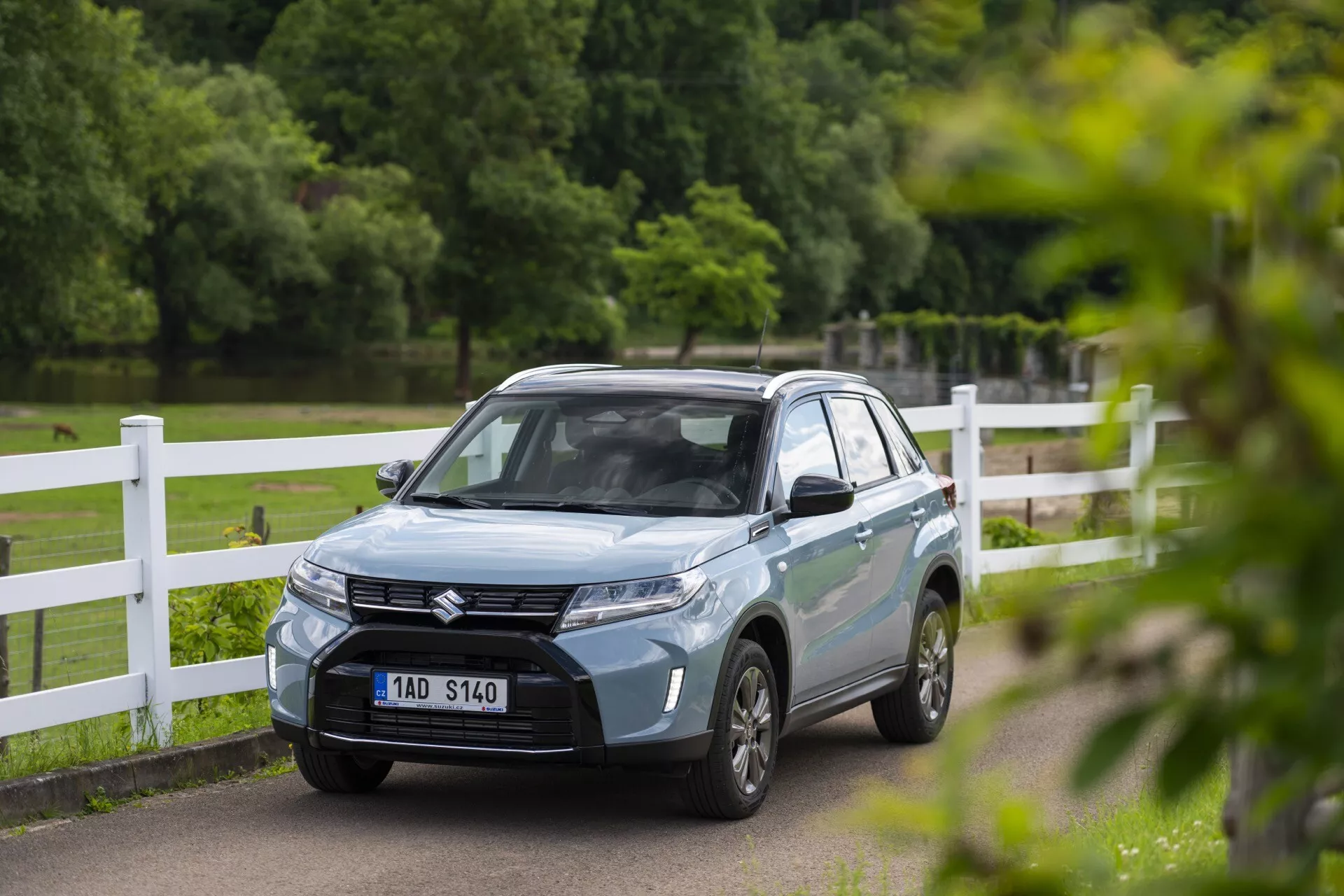 Modrý SUV Suzuki Vitara 2024 v parku nebo venkovském prostředí, zobrazený z předního tříčtvrtečního úhlu, což ukazuje jeho sofistikovanou mřížku s emblémem, ostrá LED světla, aerodynamické linie kapoty a vícepaprsková litá kola. Přední registrační značka vozidla je rozmazaná a zpětná zrcátka mohou mít integrované ukazatele směru. Fotografie zachycuje vůz s mělkou hloubkou ostrosti, zdůrazňující jeho moderní design, zatímco pozadí zůstává mírně rozmazané.